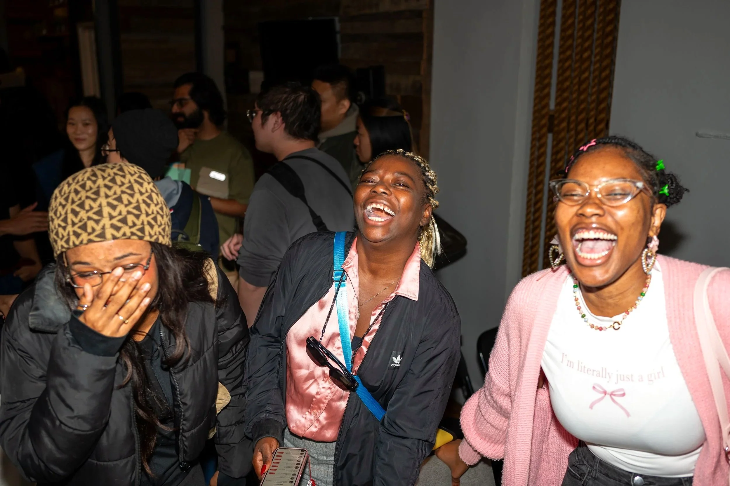 Three women laughing and smiling at a social gathering, with other people in the background.