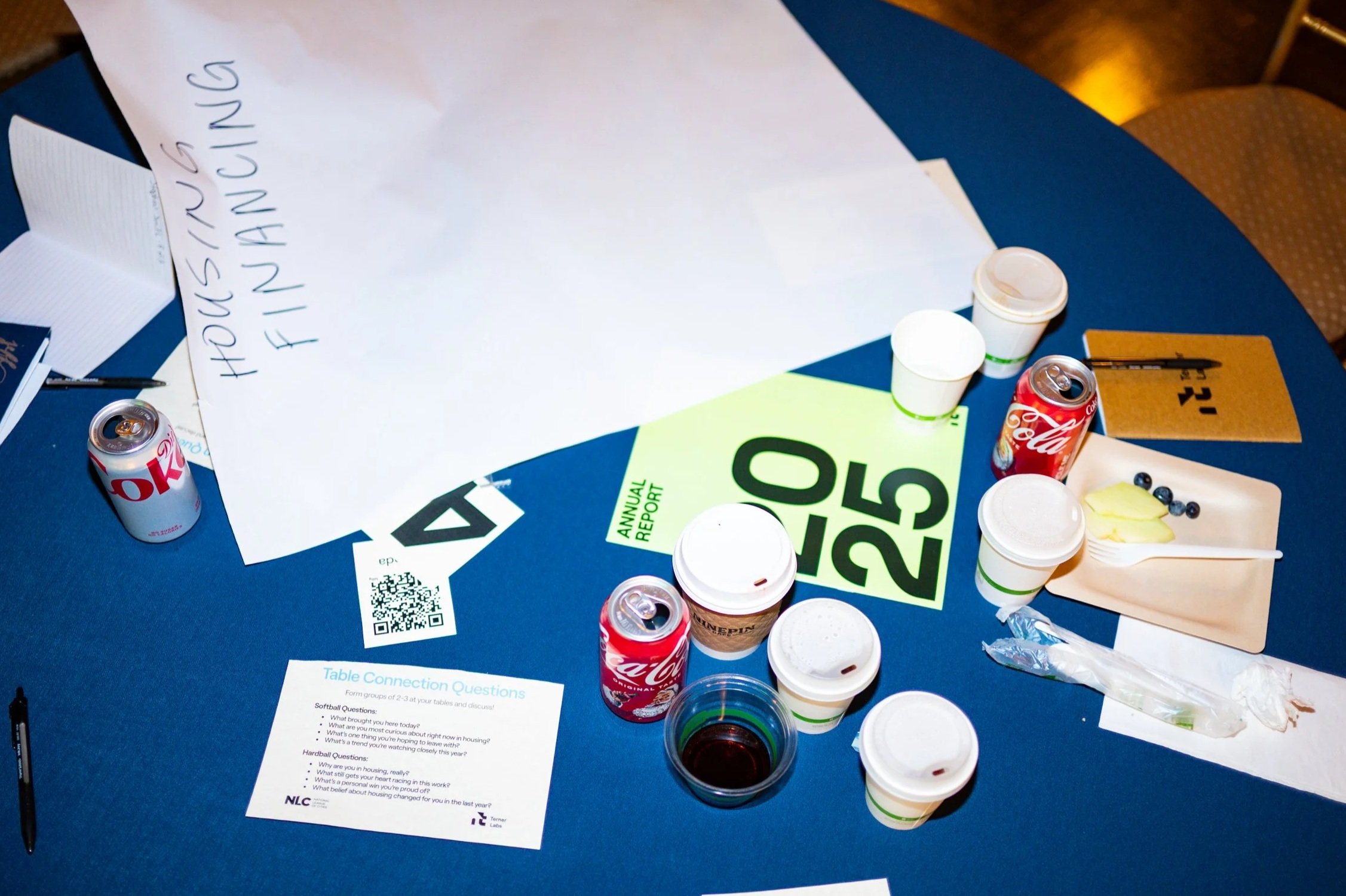 A blue table with various papers, cups, soda cans, and a plate with butter and blueberries. One large sheet of paper has '#HOUSE TOUR FINAL DINING' written on it. Other papers include a green '2025' and an 'Annual Report'.