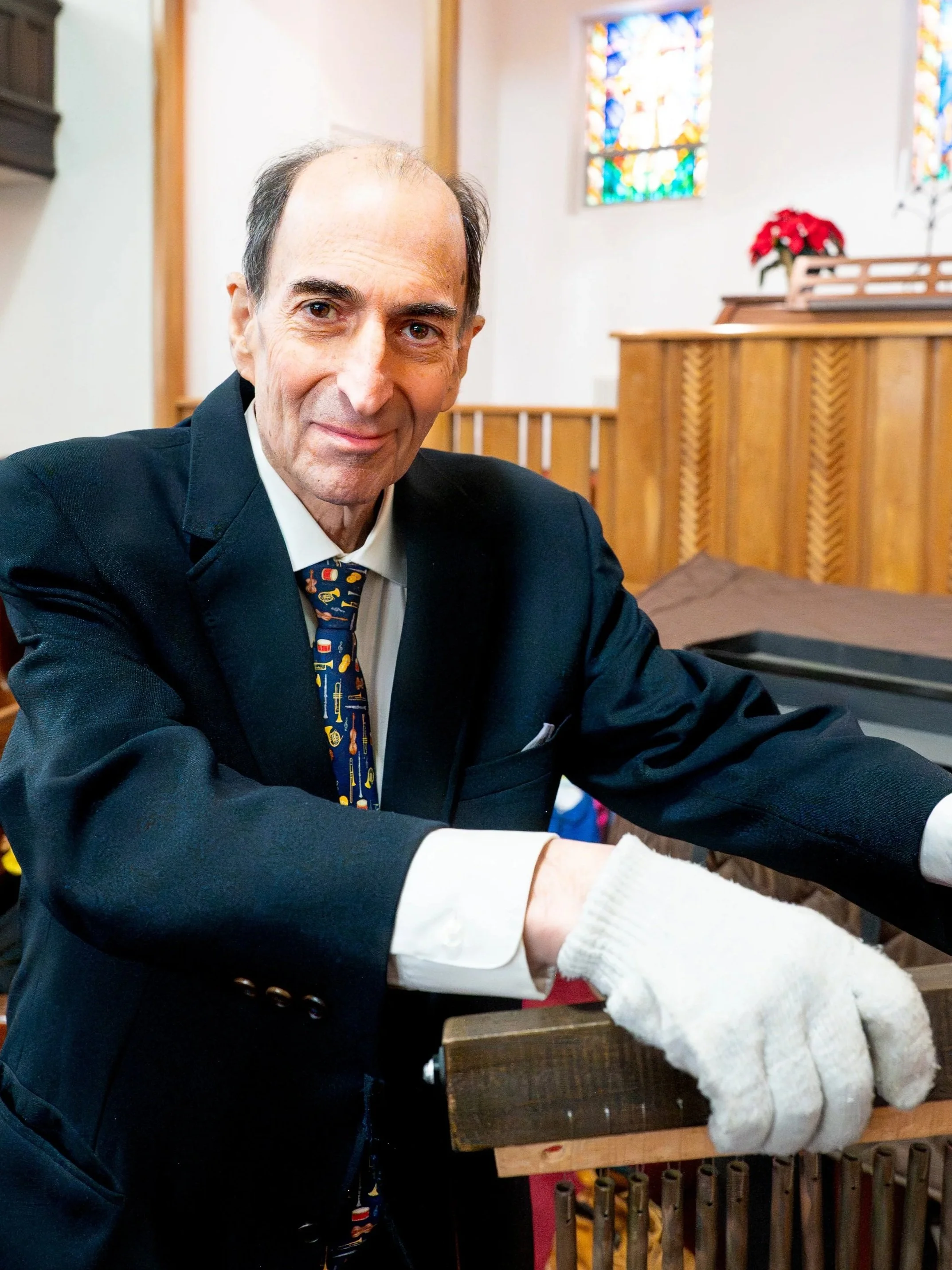 An elderly man in a black suit with a colorful tie, wearing white gloves, inside a church with stained glass windows and wooden pews.