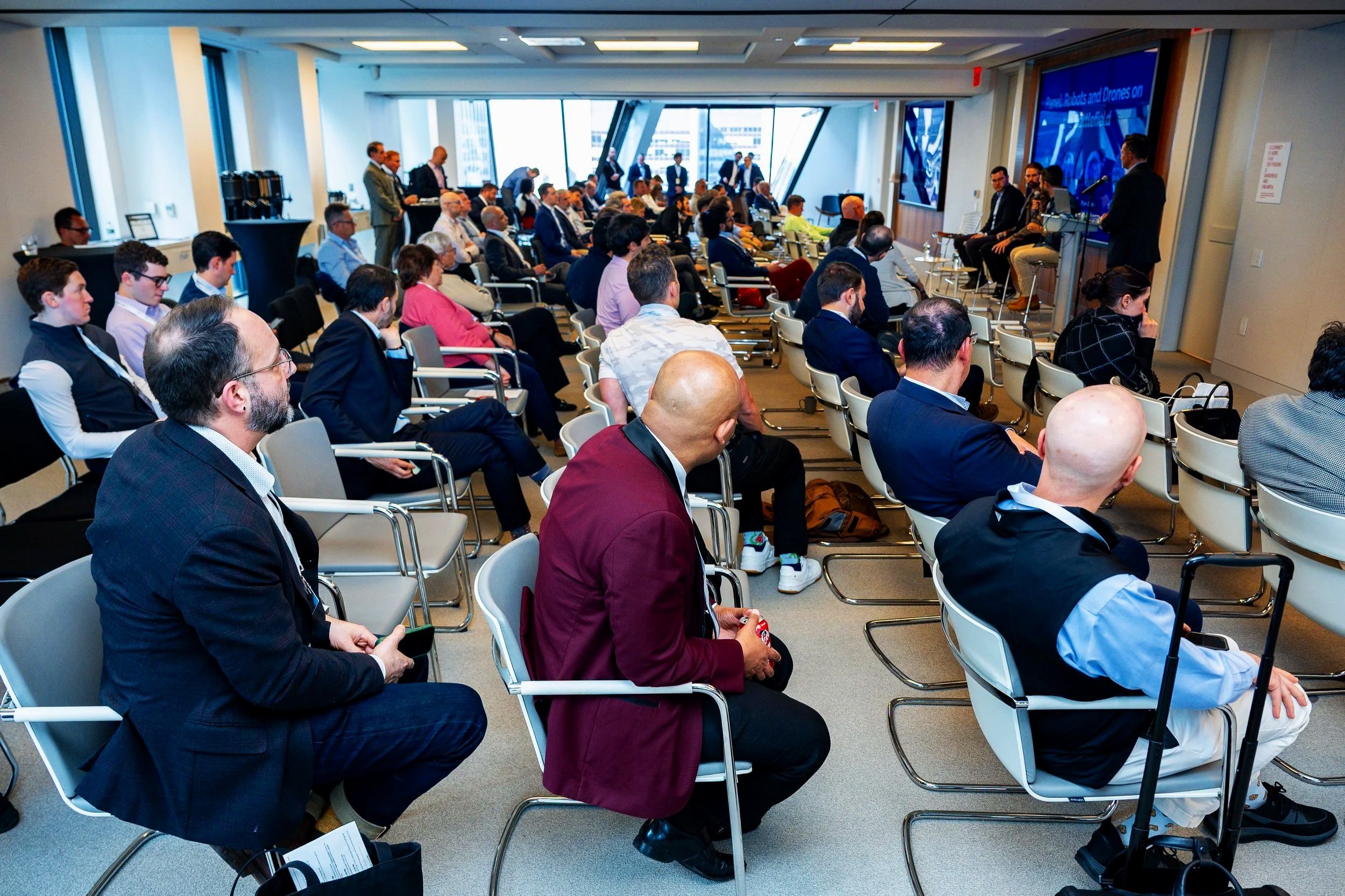 A professional conference with many attendees seated in a modern, bright room listening to a panel of speakers at the front. Large windows overlooking a cityscape are visible.