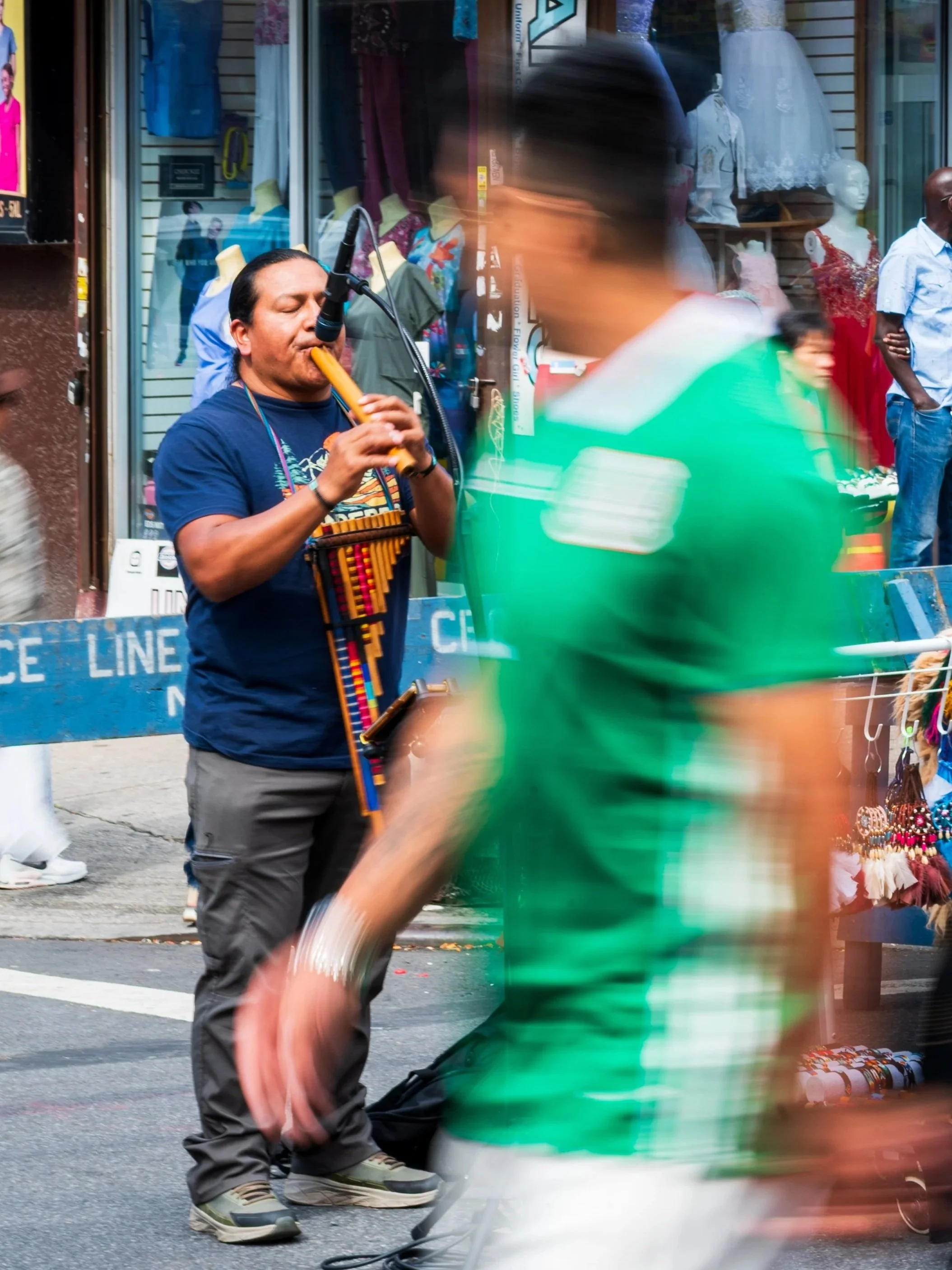 A street performer playing a flute in front of a storefront with mannequins dressed in formal attire. Pedestrians walk by, some blurred due to motion, on a busy city sidewalk. A police line cordons off part of the street.