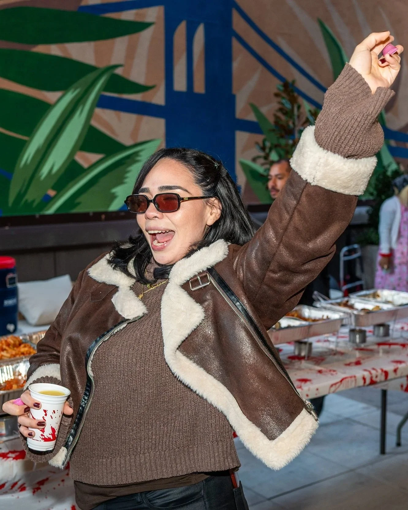 Woman wearing sunglasses and a brown jacket with cream shearling trimming cheerfully dancing or celebrating at a party with a table of food and others in the background.