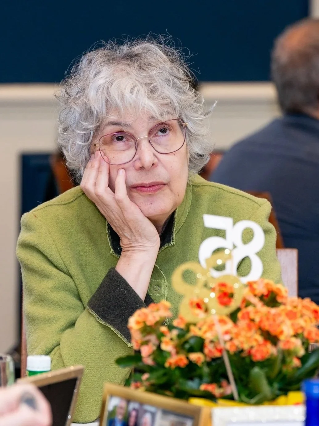 An older woman with curly gray hair and glasses resting her chin on her hand at a table decorated with flowers and framed photos, attending a social gathering or celebration.