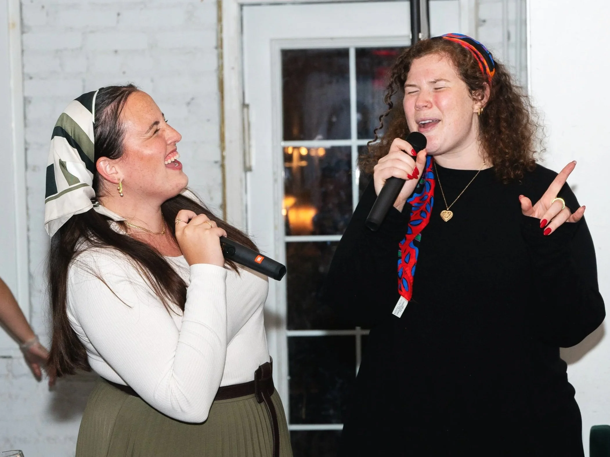 Two women singing karaoke and having fun, one with a microphone and another with a microphone, indoors with a white wall and window in the background.