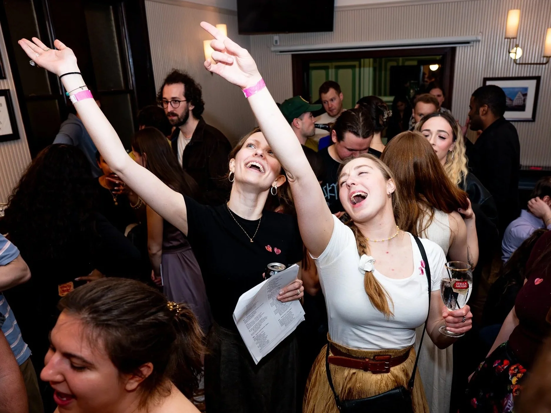 A lively group of young adults is gathered at a party, dancing and enjoying drinks. The setting appears to be a cozy indoor venue with framed pictures on the wall and warm lighting.
