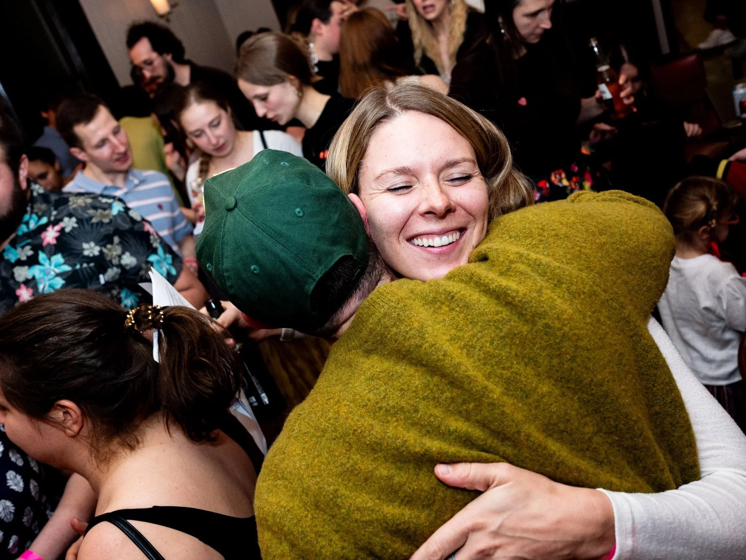 People hugging closely, with a smiling woman in the center. The scene appears to be at a social gathering or celebration.