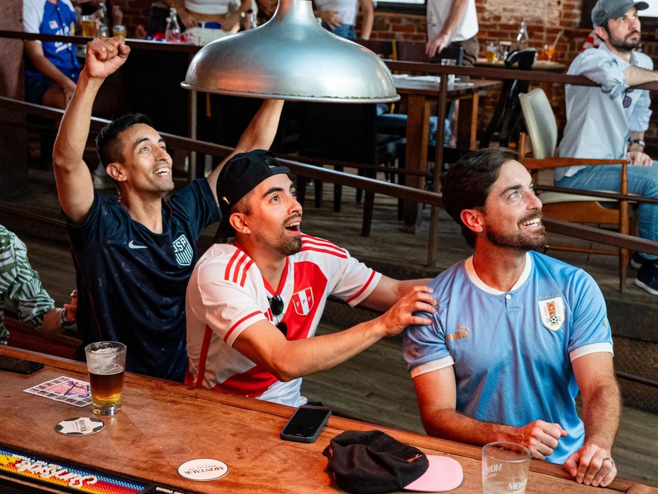 Group of people sitting at a wooden table in a restaurant or bar, enjoying drinks and socializing. Some are smiling and looking upward, while one person is lifting a large metal lamp or cover above their head.