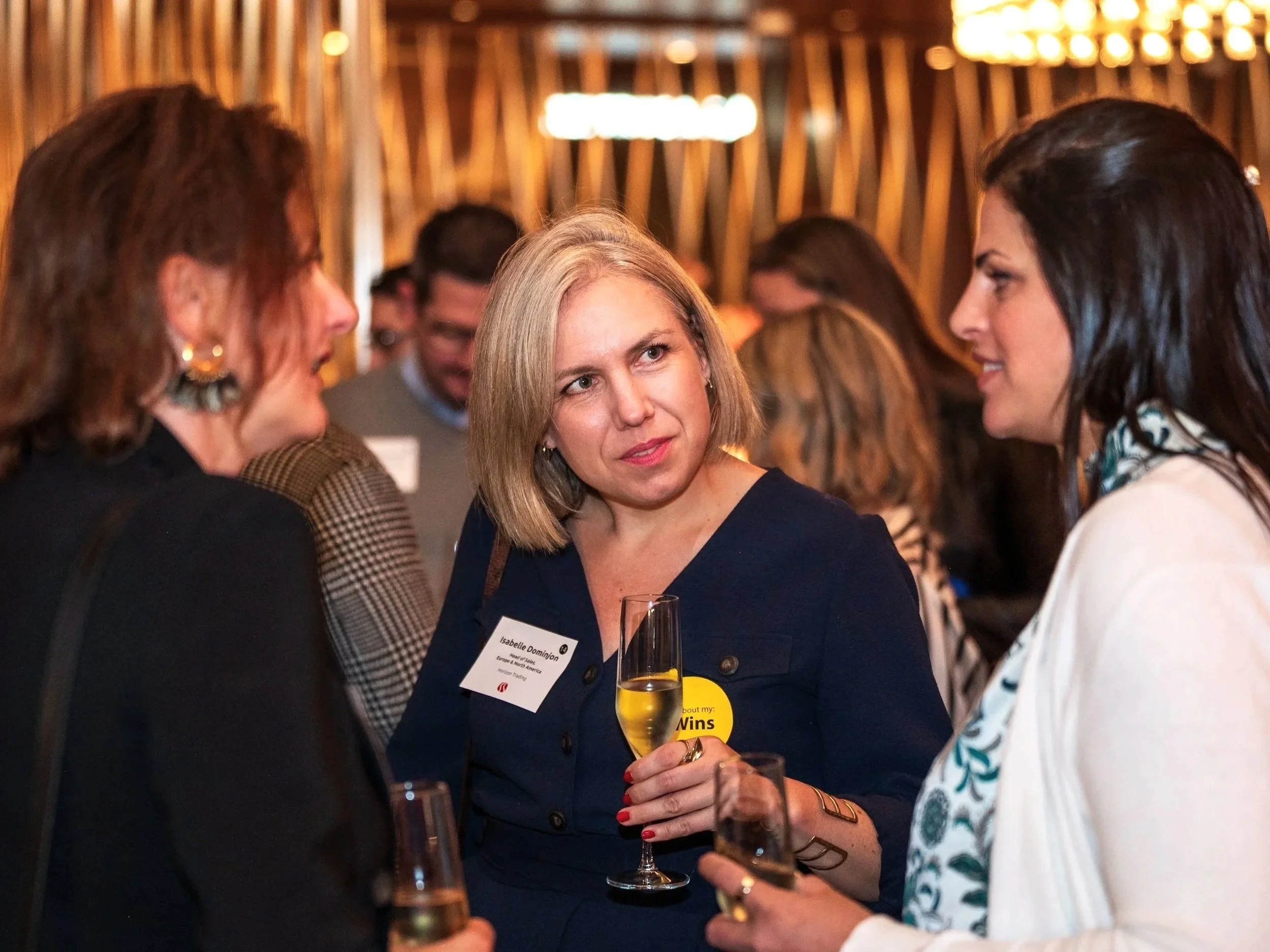 Women at a networking event conversing, some holding wine glasses, with warm lighting and wooden decor in the background.