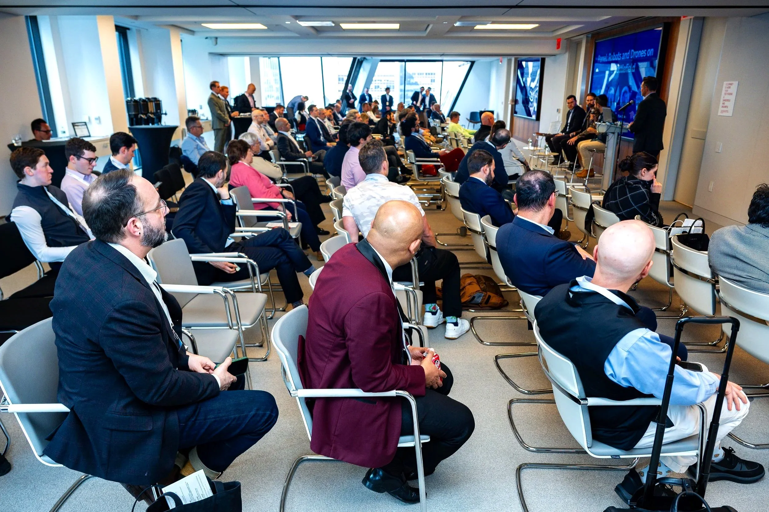 A conference room filled with attendees sitting in chairs and listening to a panel of speakers at the front, with a large digital screen displaying a presentation.