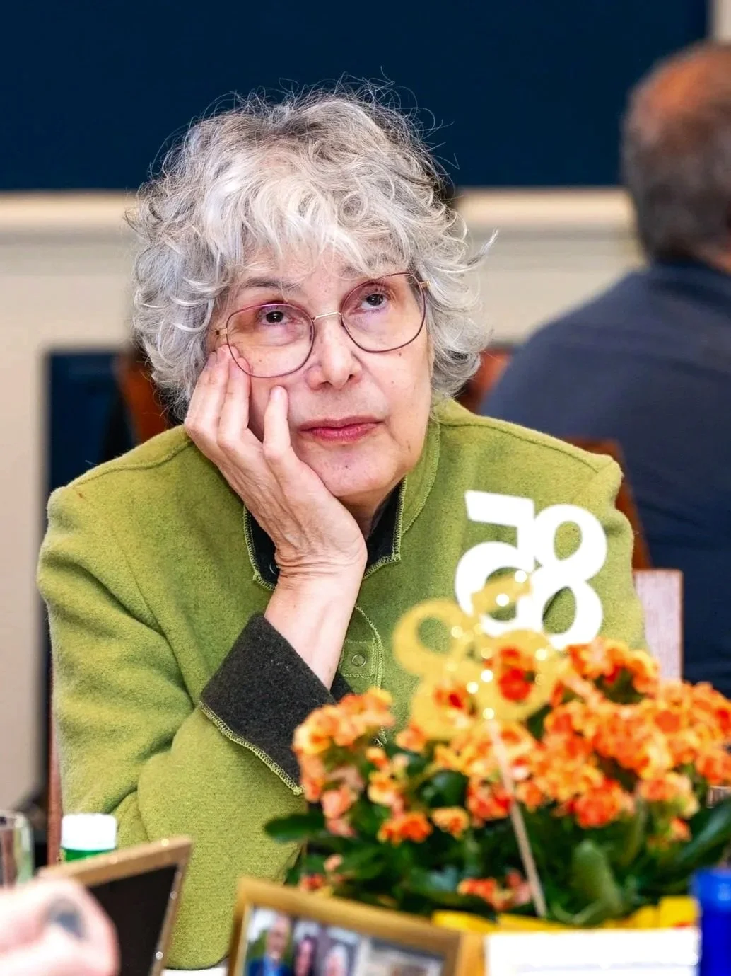 An older woman with curly gray hair and glasses resting her chin on her hand at a table decorated with flowers and framed photos, attending a social gathering or celebration.