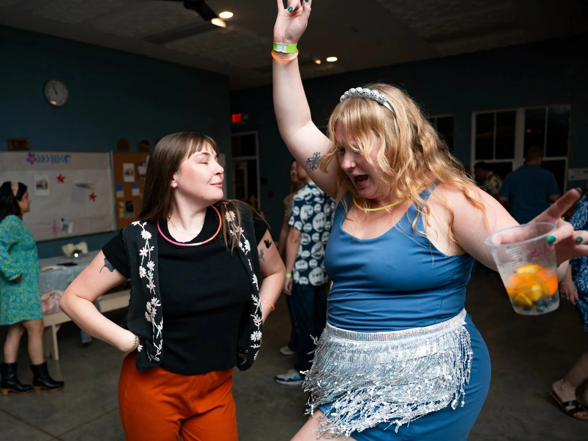 Two women are dancing and enjoying themselves at an indoor party. One woman with blonde, curly hair is wearing a shiny, beaded skirt and a blue top, raising one arm while holding a drink. The other woman with straight, brown hair is wearing a black s