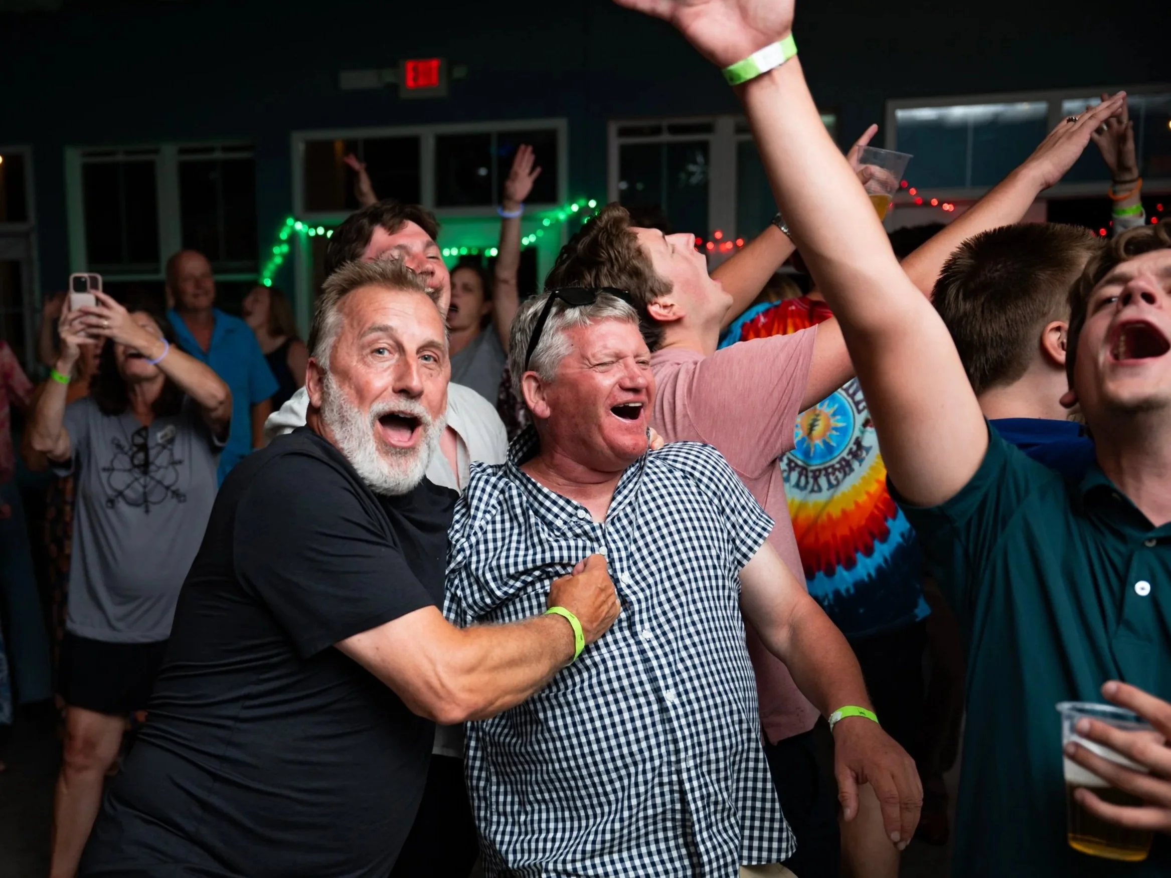 A group of people at a concert or event, appear to be enjoying and dancing, with some raising their hands and others taking photos or videos, in a dark indoor setting with colorful lights.