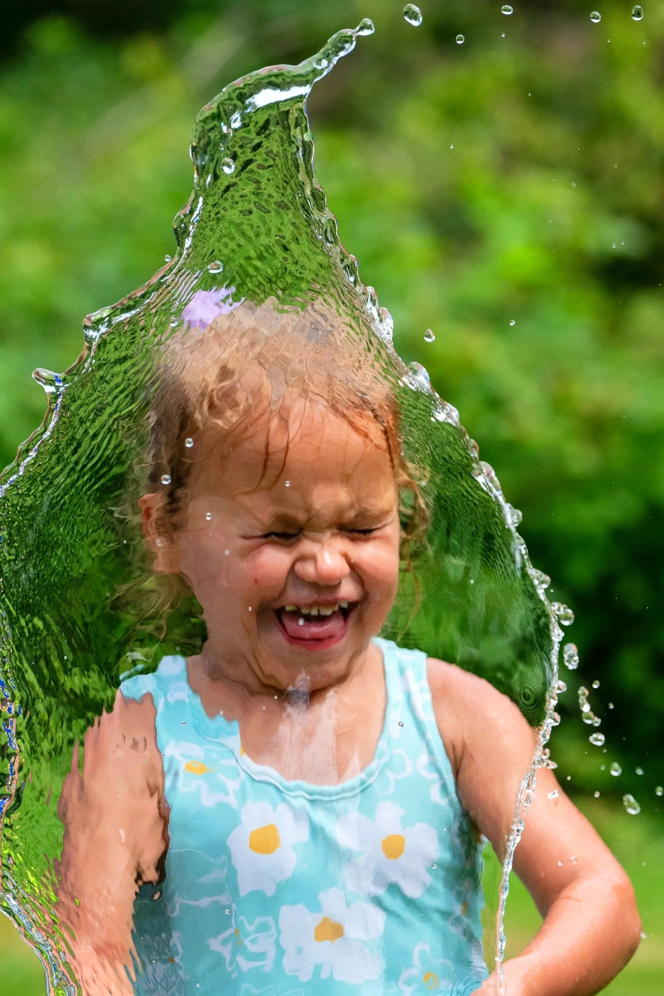 Young girl smiling and playing with water outdoors, splashing water over her face, wearing a sleeveless dress with a floral pattern, holding a green bucket.