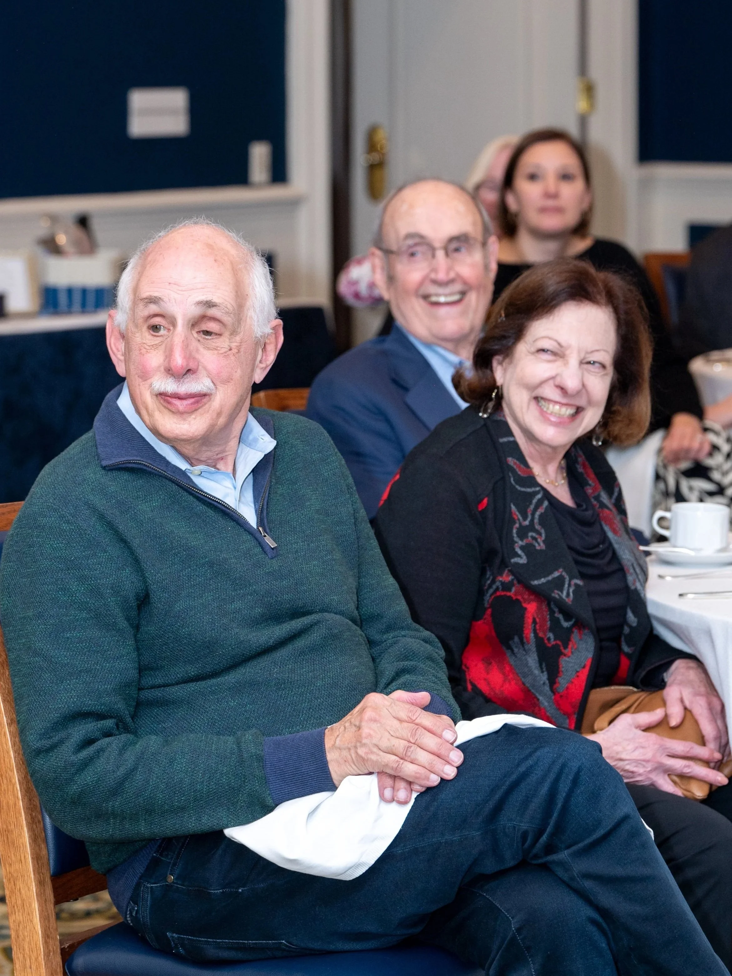 Group of elderly people sitting at a formal event, smiling and laughing, with a woman in the foreground wearing a colorful jacket and a man in a sweater beside her.