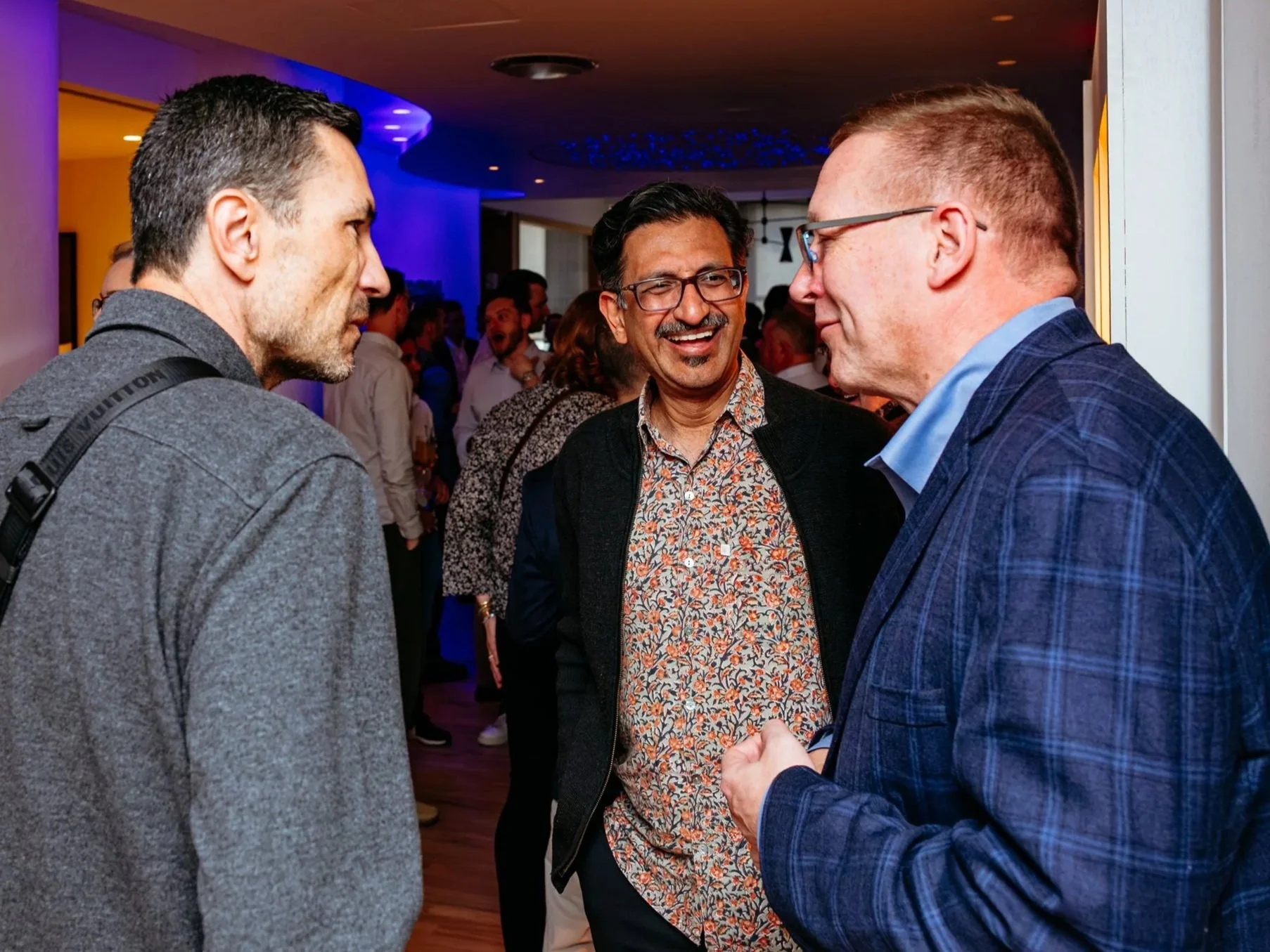 Three men are in conversation at a social gathering indoors, with other guests in the background. They are dressed in business casual attire.