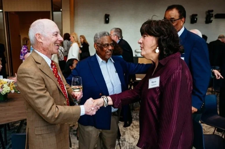People at a networking event, including three prominent individuals engaged in conversation and handshaking, with others mingling in the background.