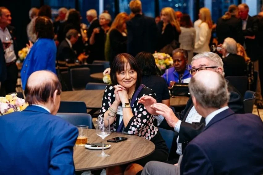 People socializing at an indoor event, some sitting at tables with flowers, others standing and mingling in background.