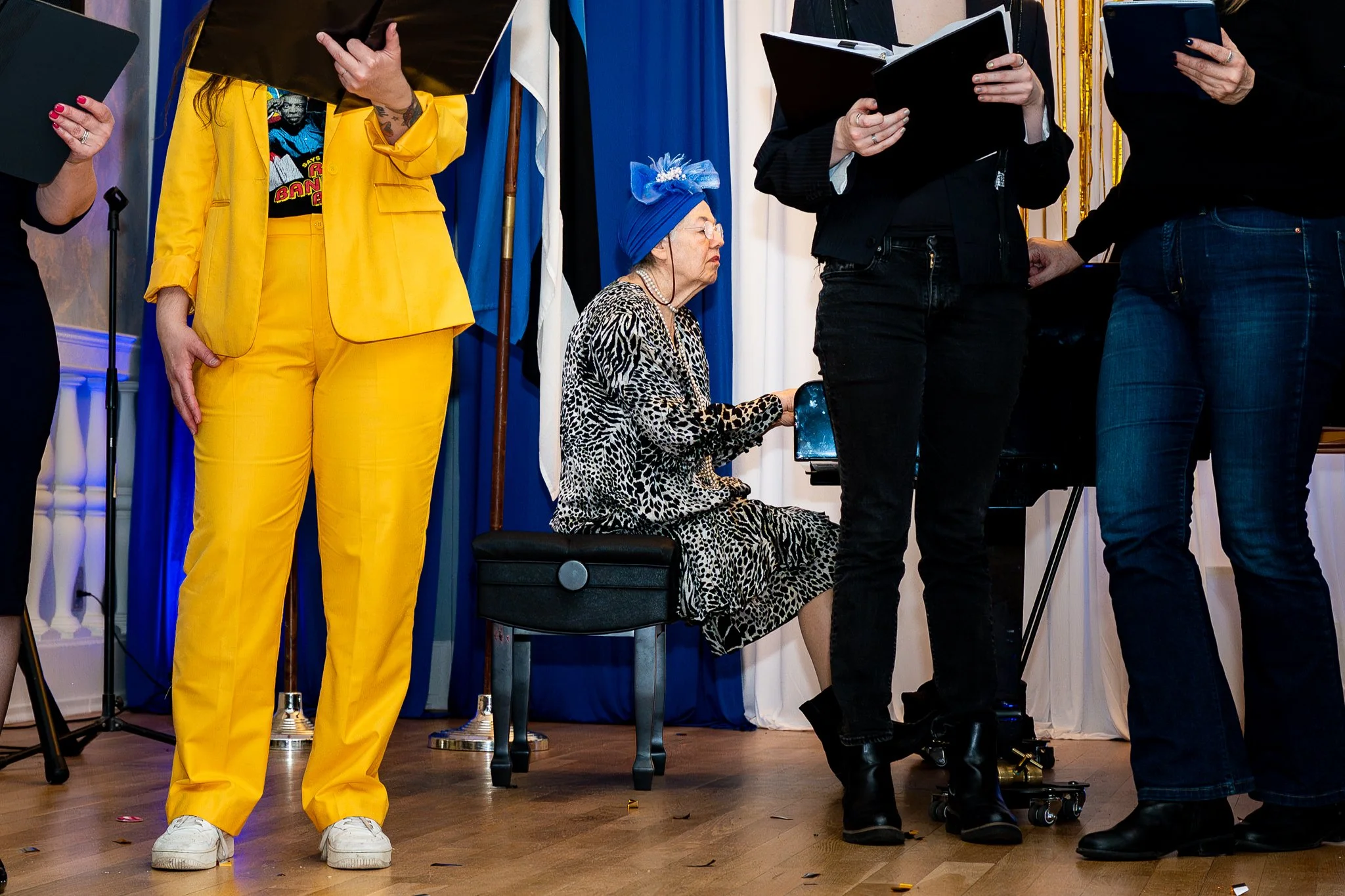 Group of women standing and sitting during an event, with one woman seated at a piano wearing a leopard print dress and a blue headscarf, others holding notebooks or tablets, in a room with royal blue curtains and gold accents.