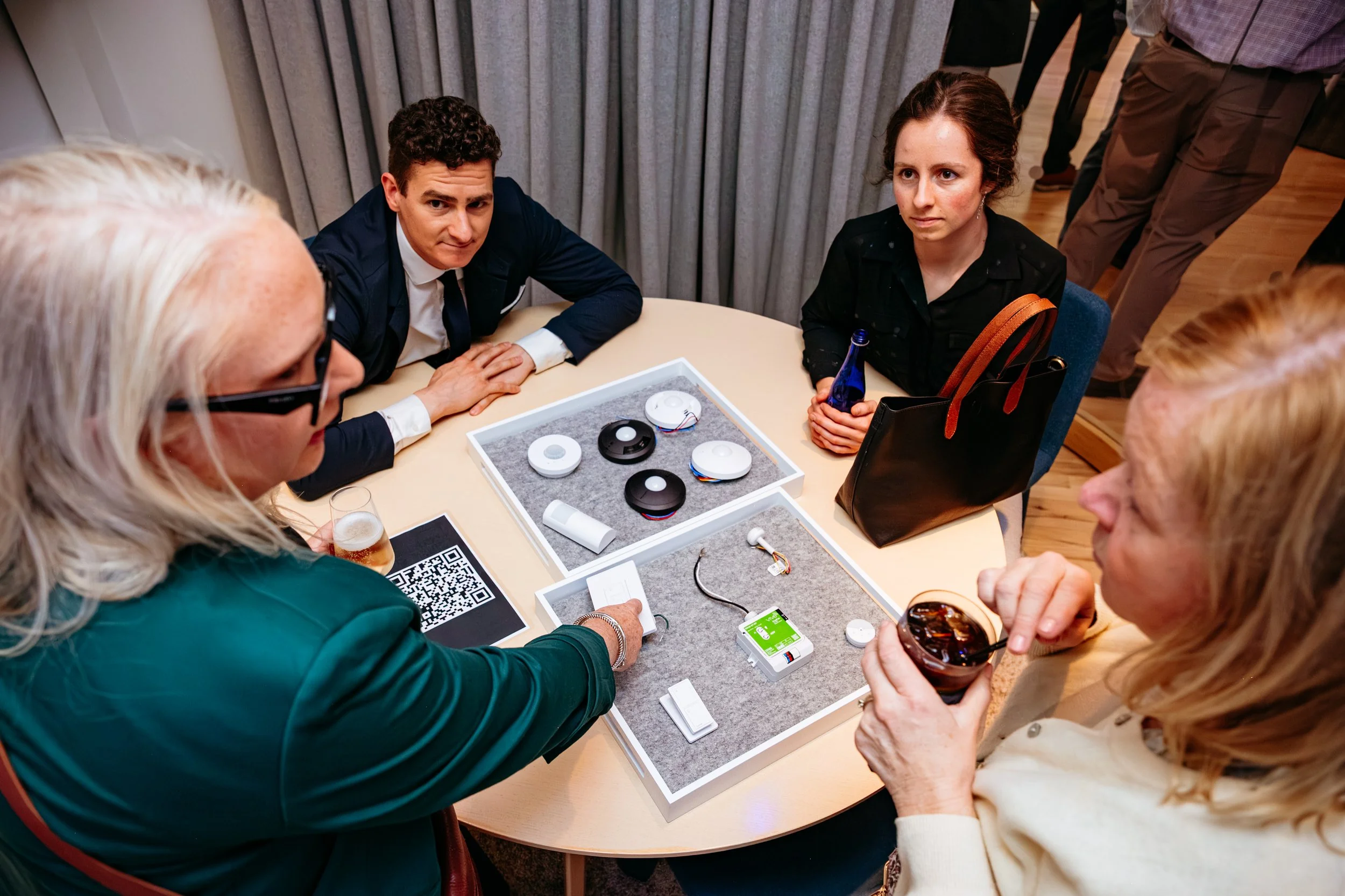 Group of people sitting around a table examining smart home devices and electronic components, with some people holding drinks.