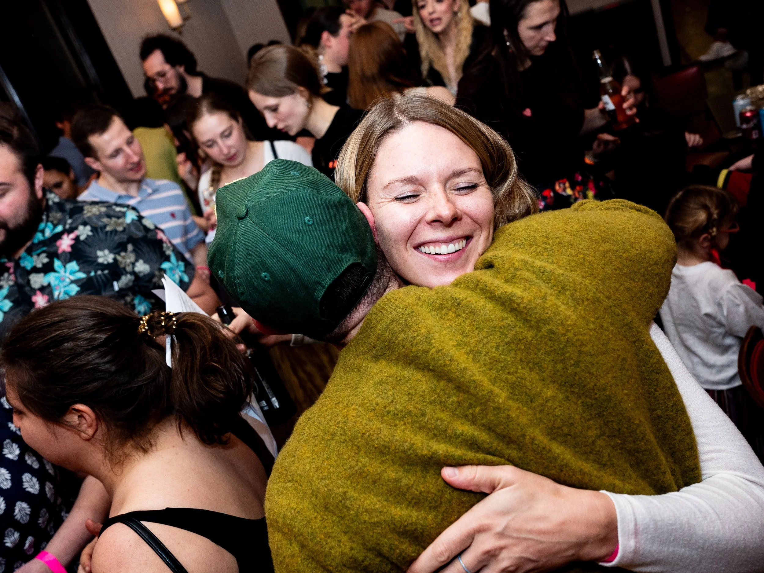 People hugging closely, with a smiling woman in the center. The scene appears to be at a social gathering or celebration.