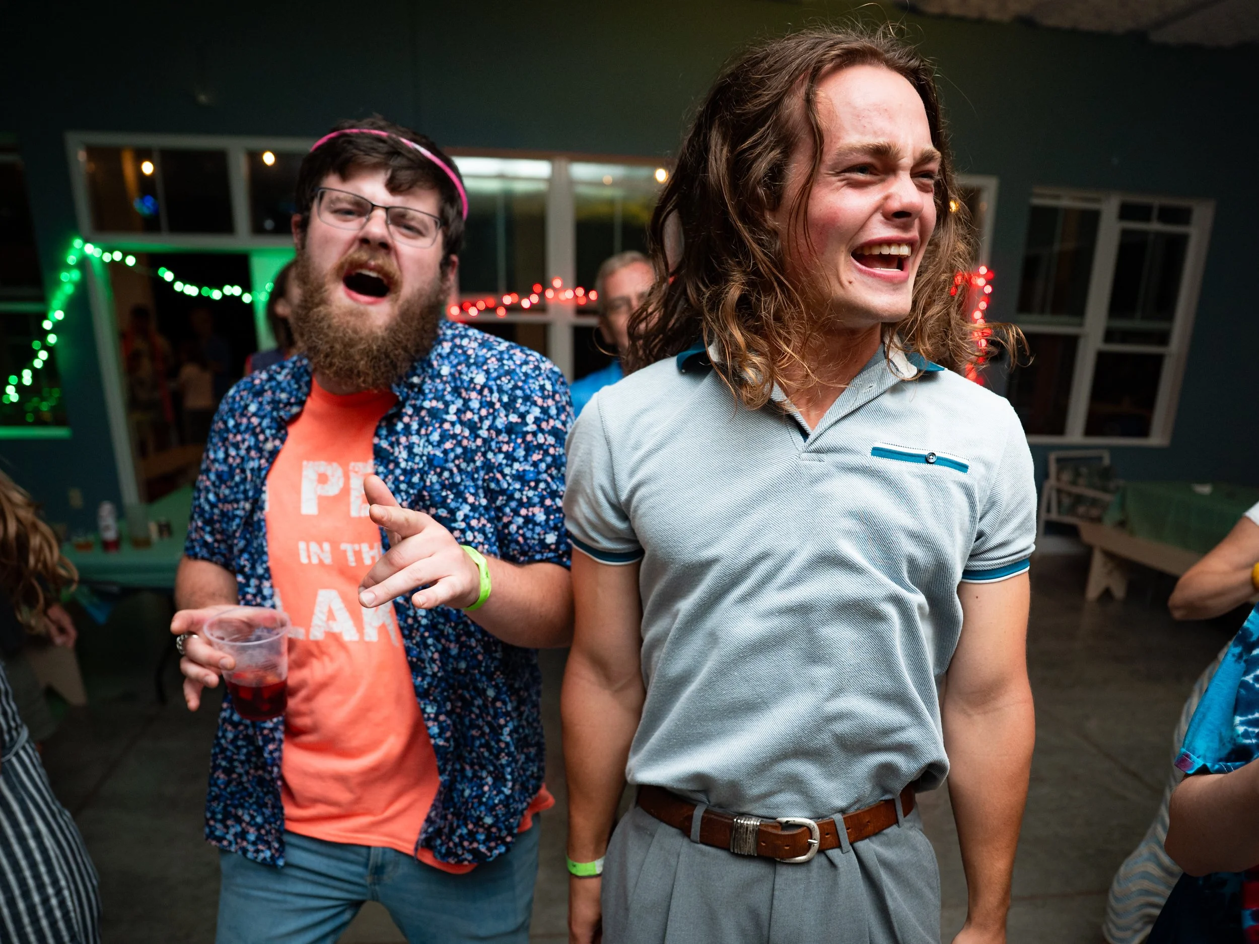 Two men are central in the image, smiling and appearing to enjoy a moment at a party or social gathering. The man in the foreground has long, curly brown hair, is wearing a light gray polo shirt and a brown belt. The man behind him has dark hair, gla