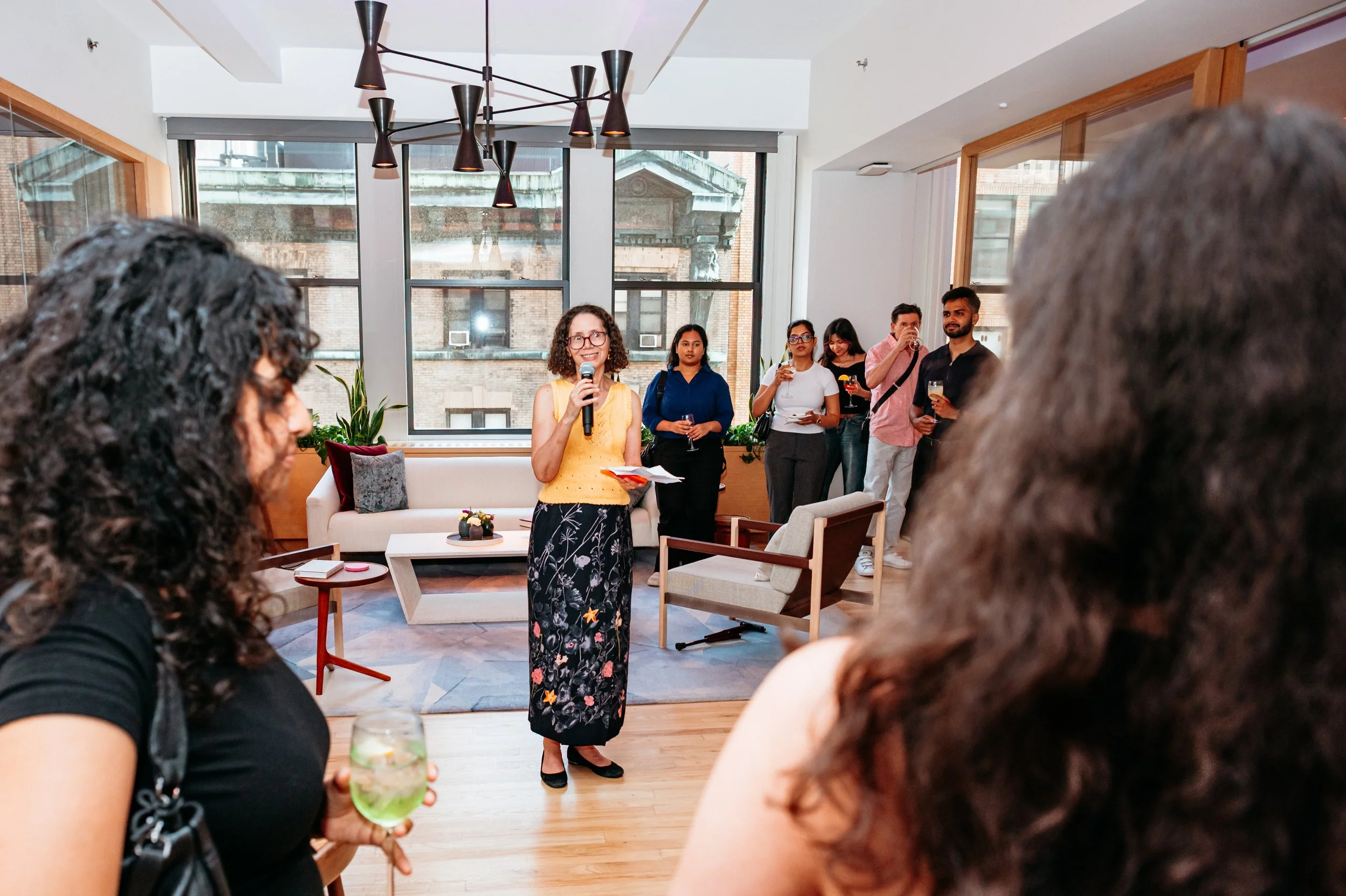 A woman with glasses in a yellow sleeveless top and a floral skirt is speaking into a microphone at a gathering. Several people are standing nearby, some holding drinks. The setting is an indoor space with large windows, modern lighting, and a cozy s