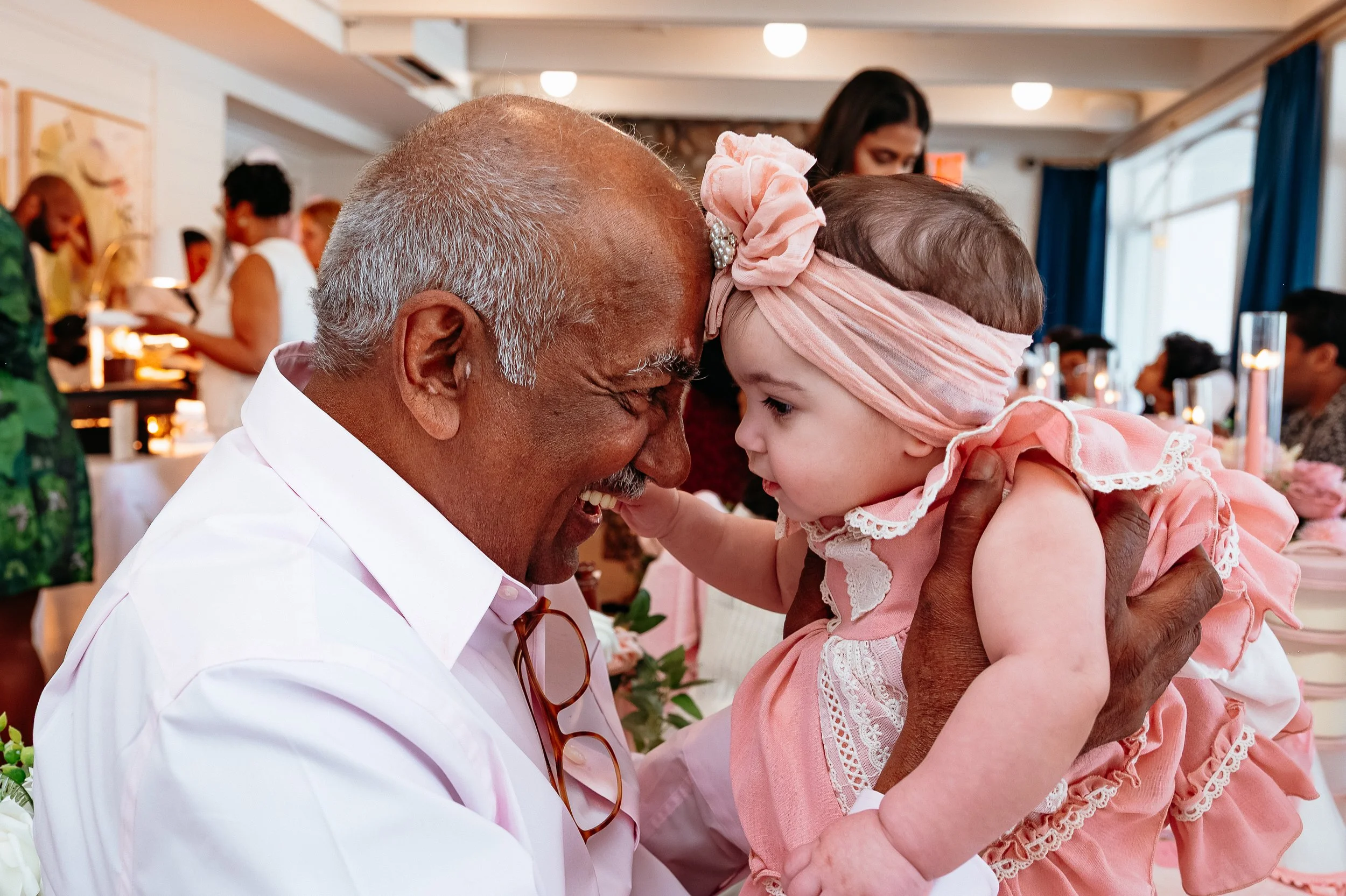 An elderly man with gray hair and a white shirt holds a young girl in a pink dress and headband in a close interaction at a social gathering.