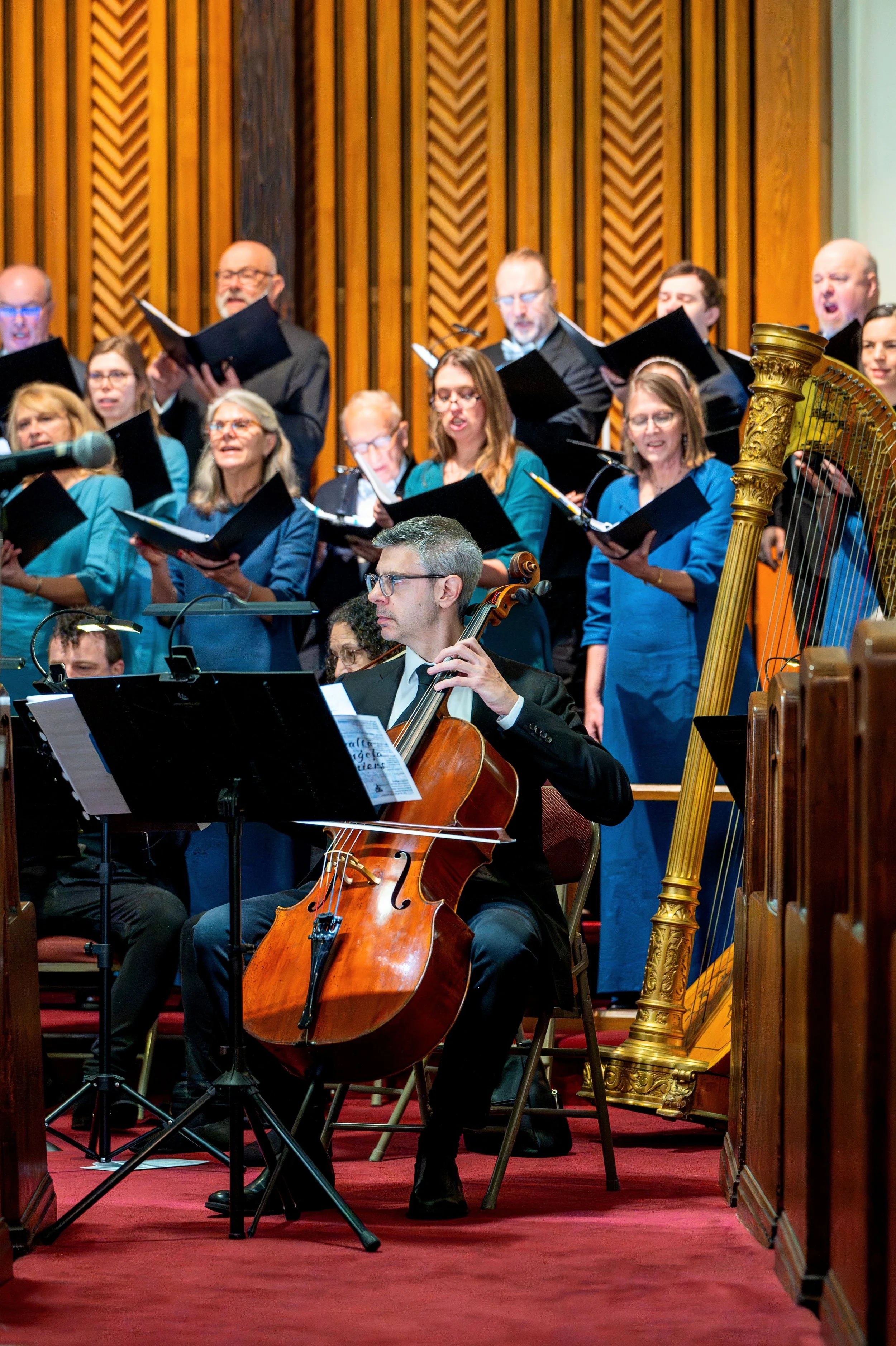 A live choir and orchestra perform on stage in a concert hall with wooden paneling. The choir members wear blue outfits, and the orchestra features a cellist, who is seated and focused on his music.