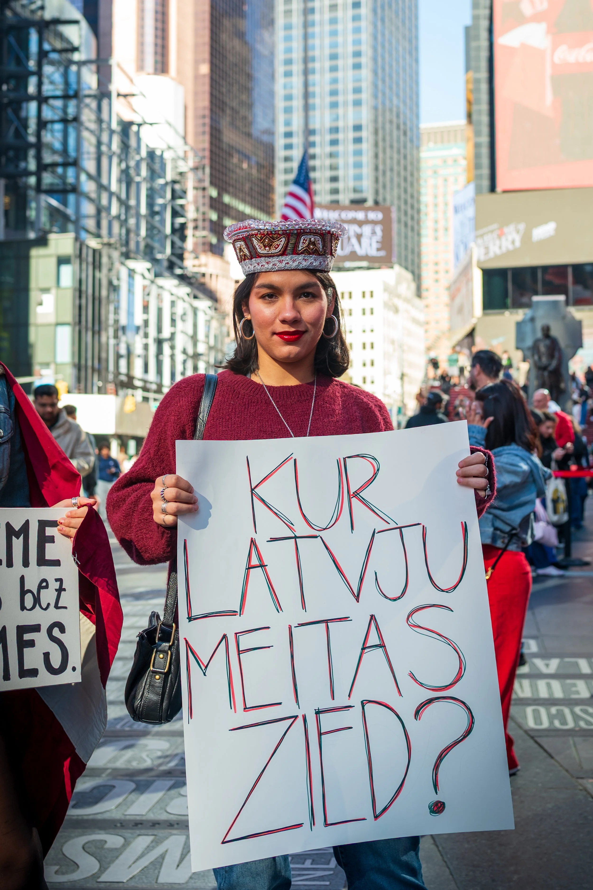 Young woman in red sweater holding a protest sign in a city with tall buildings, with other protesters nearby.