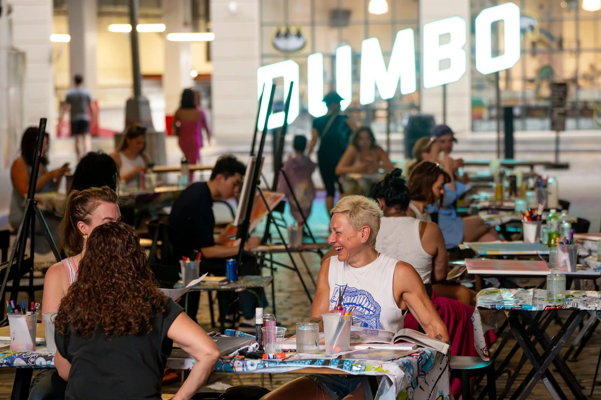 People participating in a group painting event outdoors at night, sitting at tables with art supplies, with a large illuminated sign that says 'ALIMBO' in the background.
