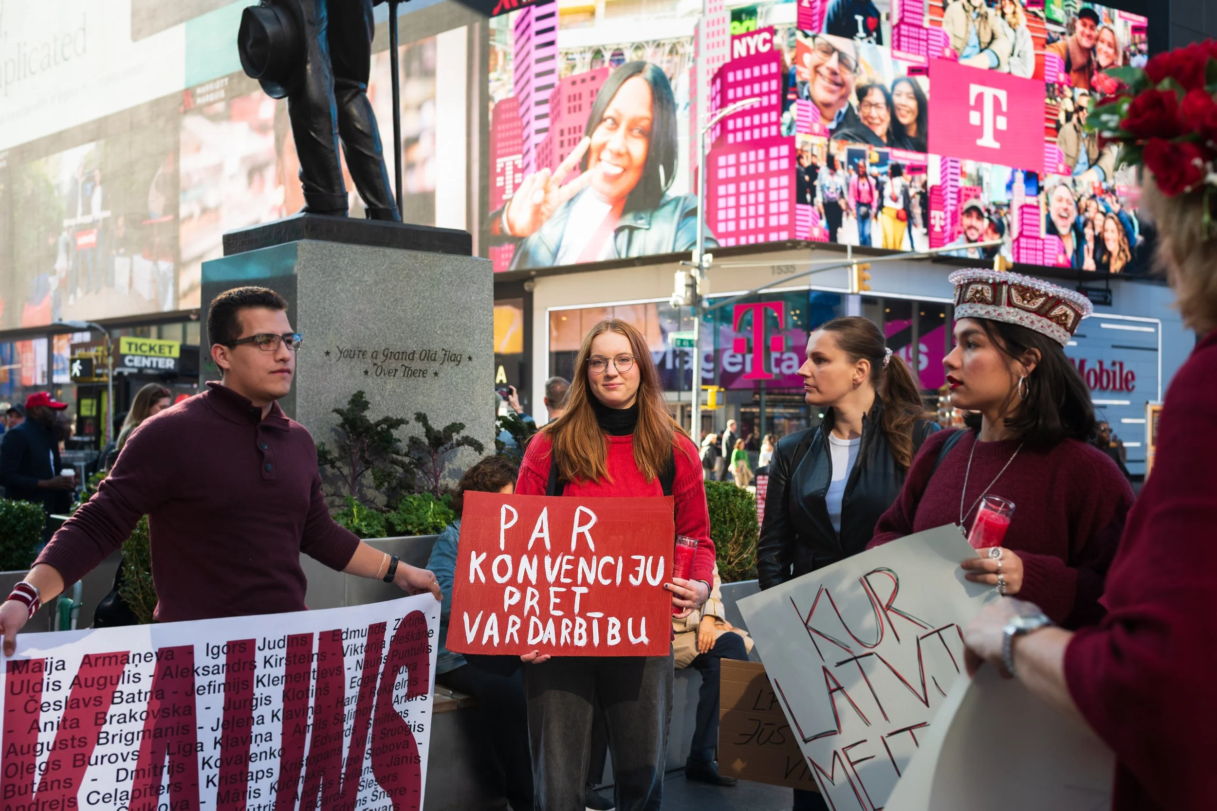 People participating in a public protest or demonstration in Times Square, New York City, holding signs with messages in Latvian. The background features digital billboards and advertisements.