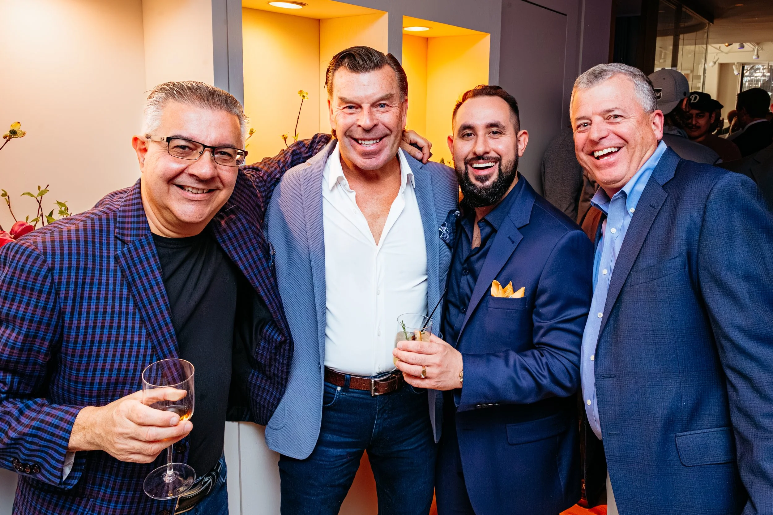 Four men in suits smiling and posing for a photo at a social event, with drinks in hand, in an indoor setting with yellow and gray walls.