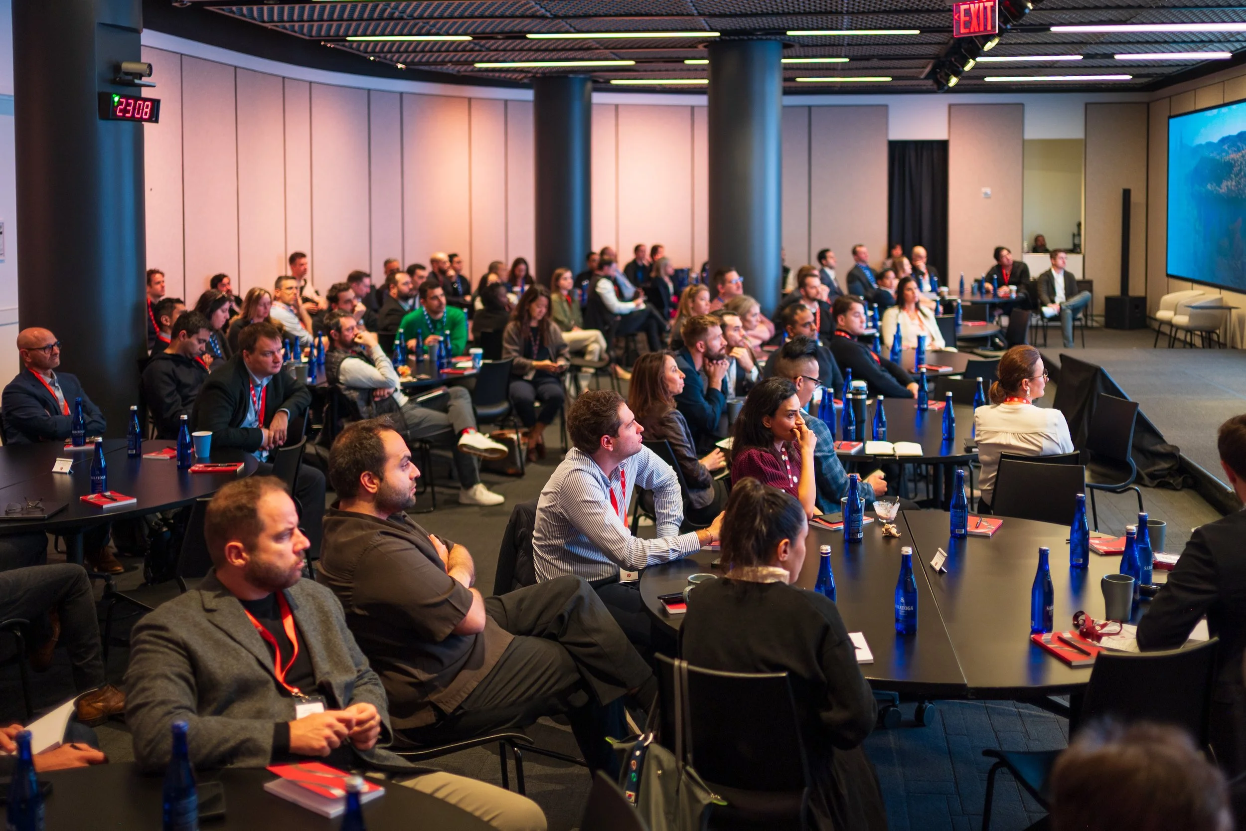 A conference room filled with attendees sitting at round tables, watching a presentation projected onto a large screen.