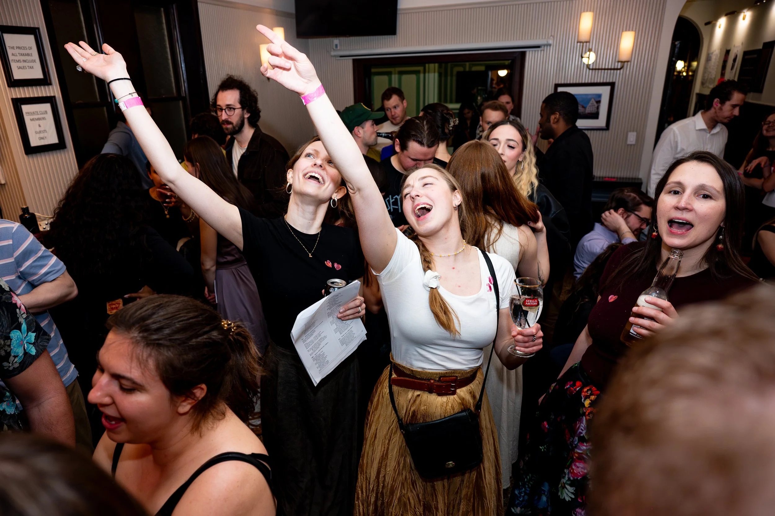 A lively group of young adults is gathered at a party, dancing and enjoying drinks. The setting appears to be a cozy indoor venue with framed pictures on the wall and warm lighting.