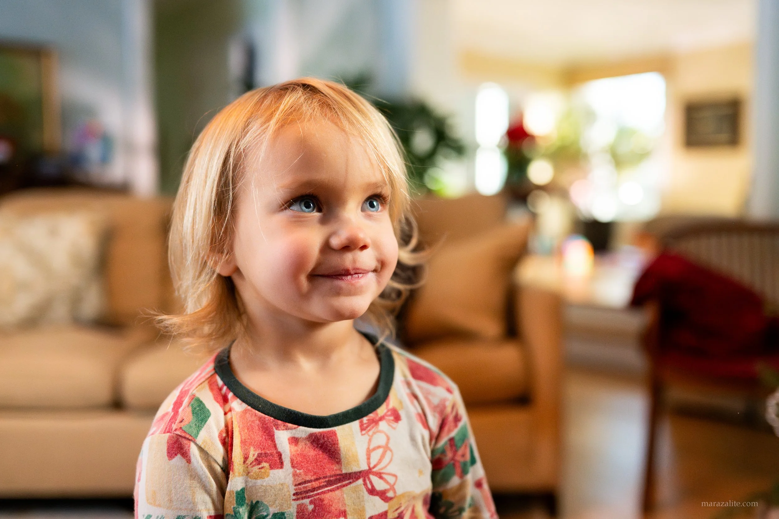 A young girl with blonde hair and blue eyes looking upwards, wearing a colorful patterned shirt, standing inside a house with blurred background of furniture and natural light.