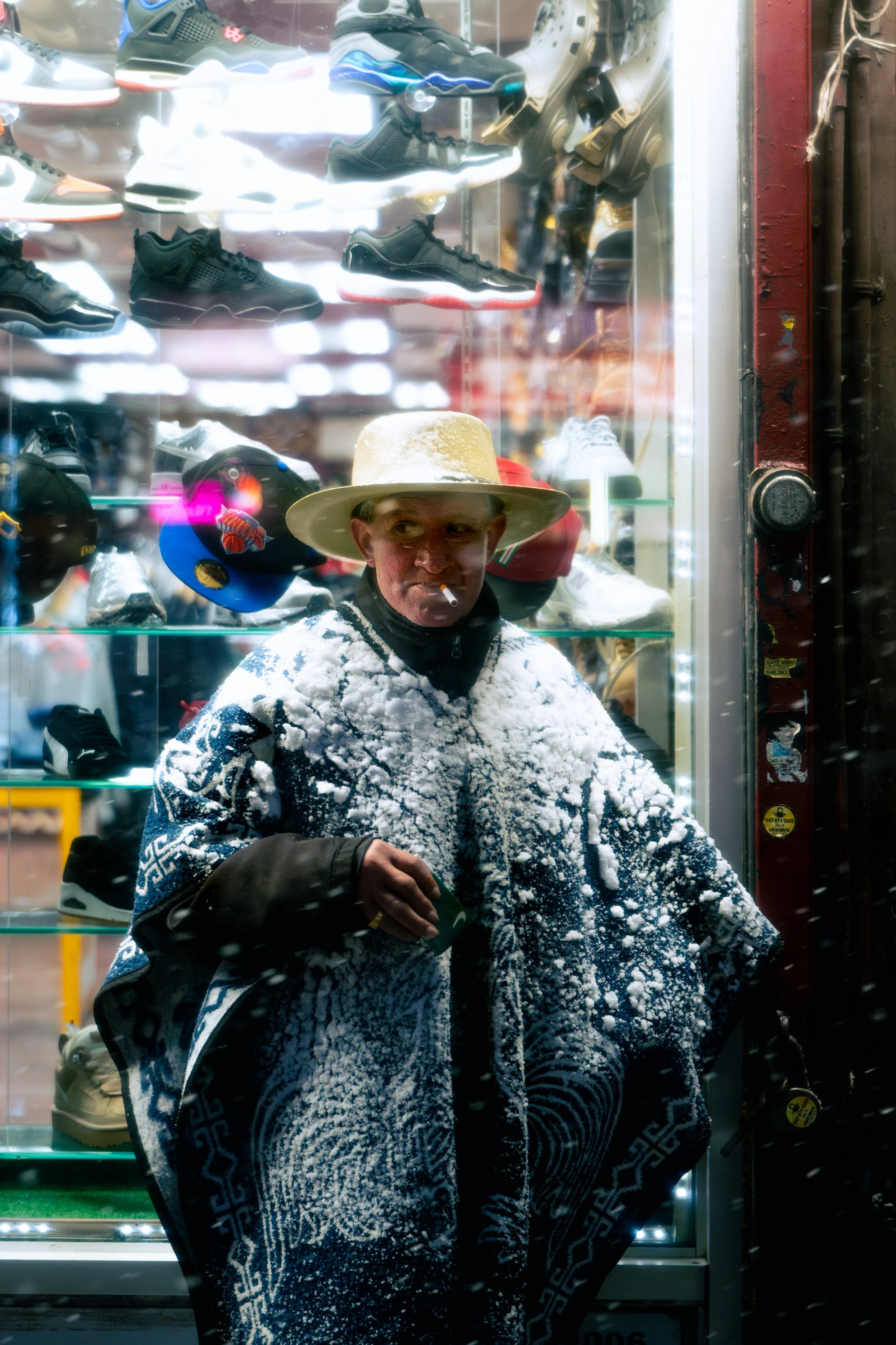 A man standing behind a glass display case of shoes and helmets, wearing a large straw hat and a blue patterned poncho, with snow on his clothing and a cigarette in his mouth.