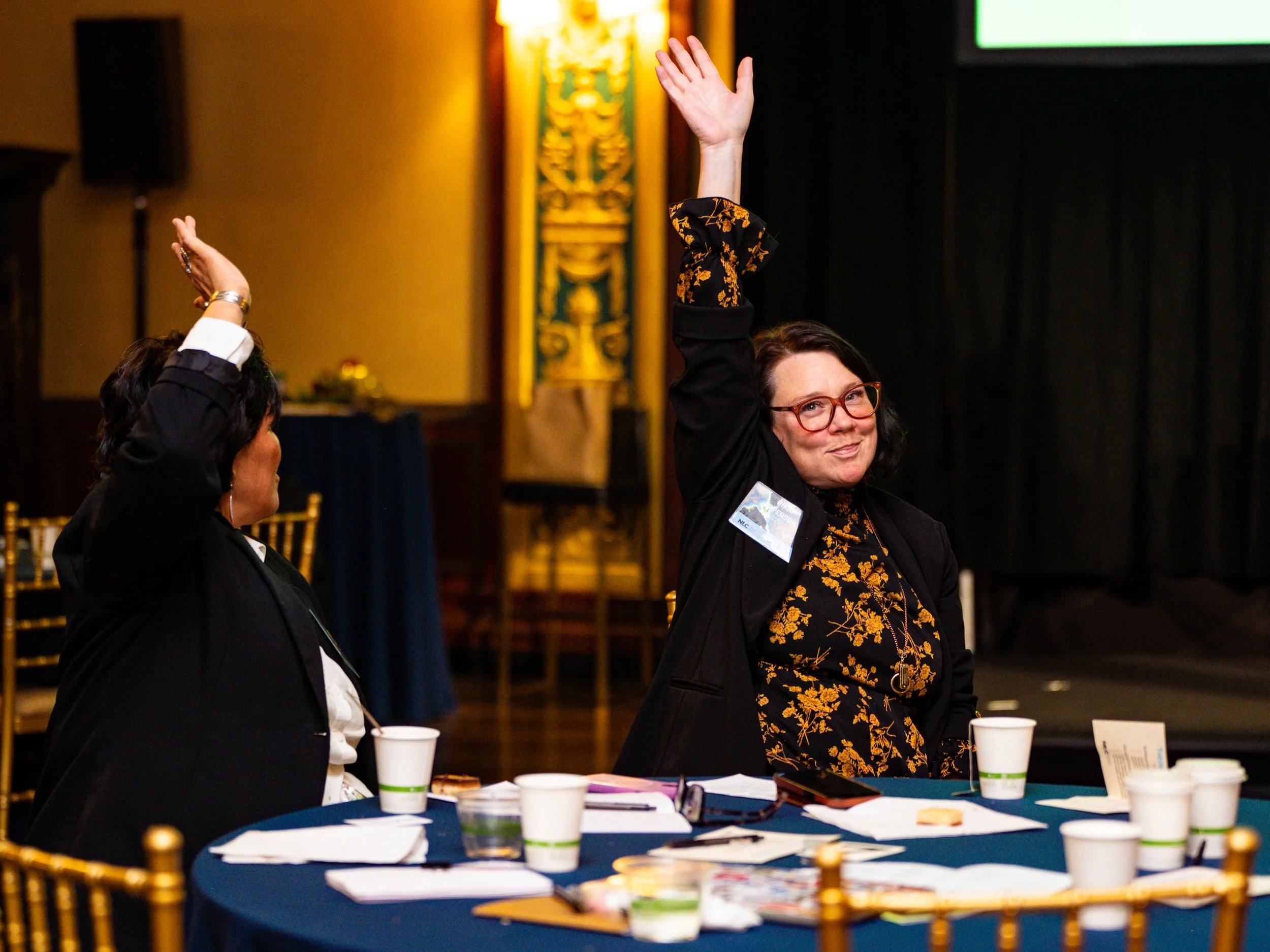 Two women sitting at a round table in a conference room, raising their hands. The woman on the right is wearing glasses and a black blazer with a patterned black and gold top underneath. The woman on the left has short dark hair and is also raising h