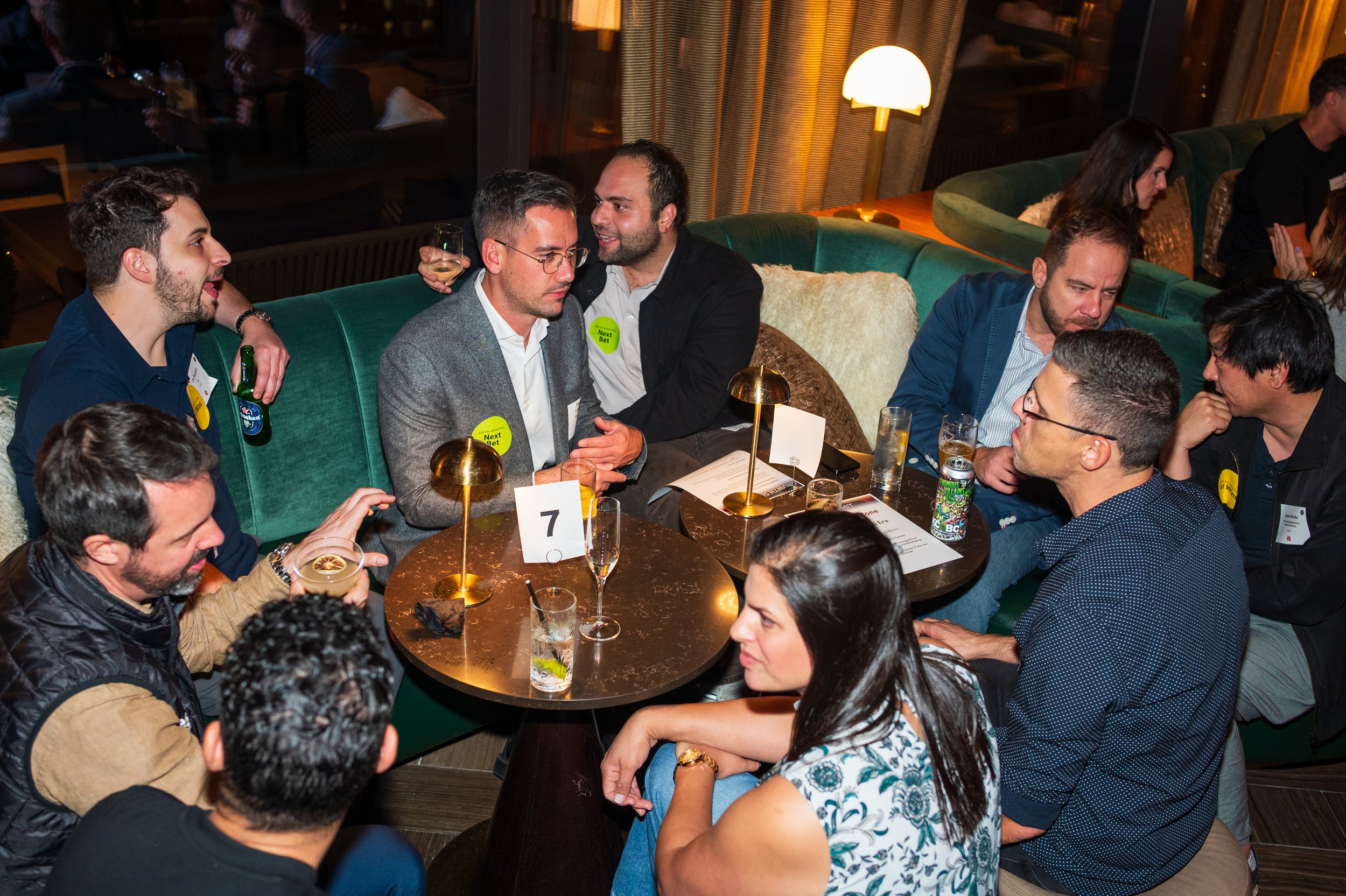 People sitting at a round table in a dimly lit restaurant or bar, engaged in conversation, some holding drinks, with a mix of men and women, some wearing name tags or stickers, and a small table number sign.