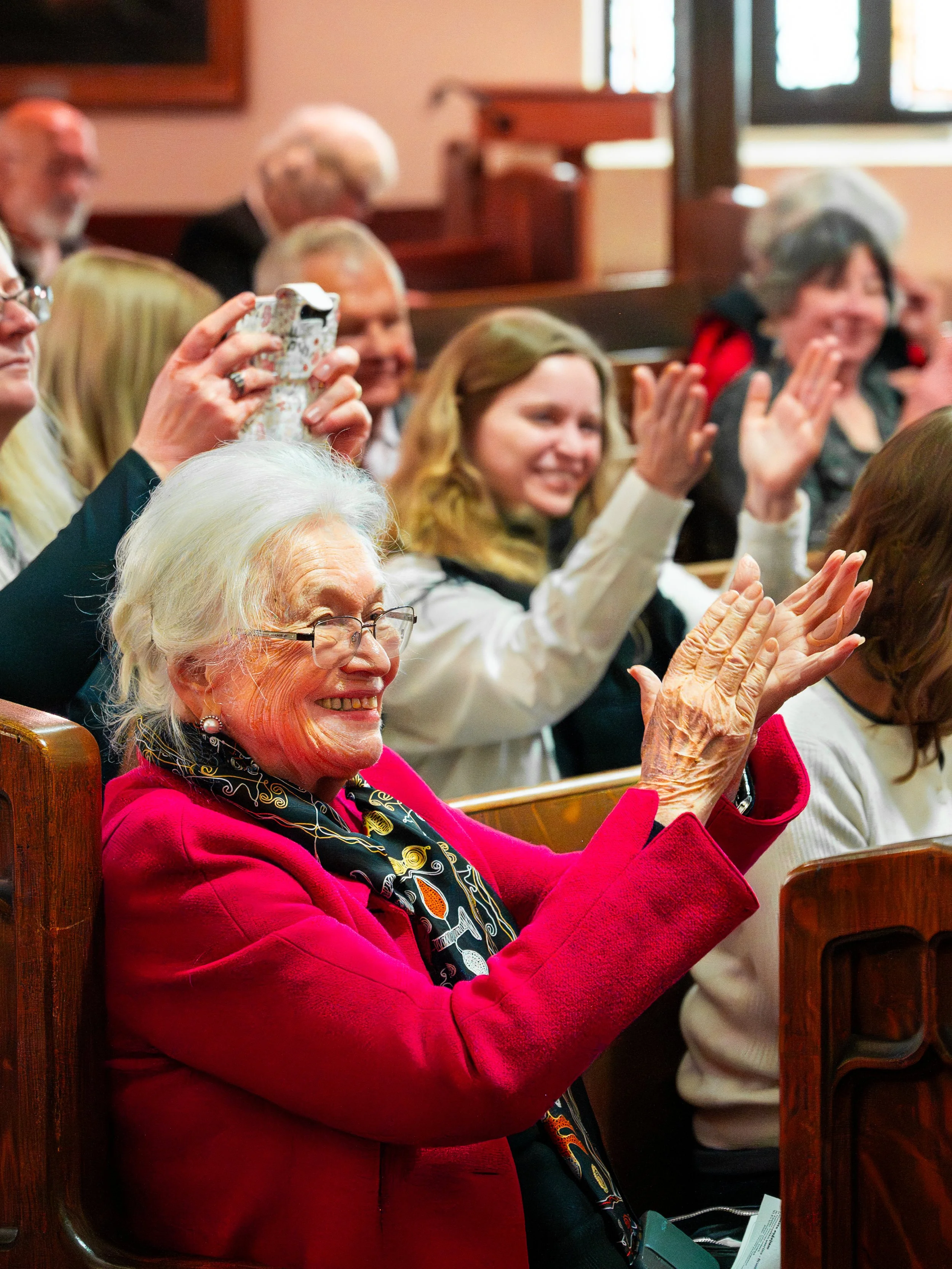 Elderly woman with white hair, wearing glasses, a pink coat, and a black patterned scarf, smiling and clapping in a crowd at an indoor event.
