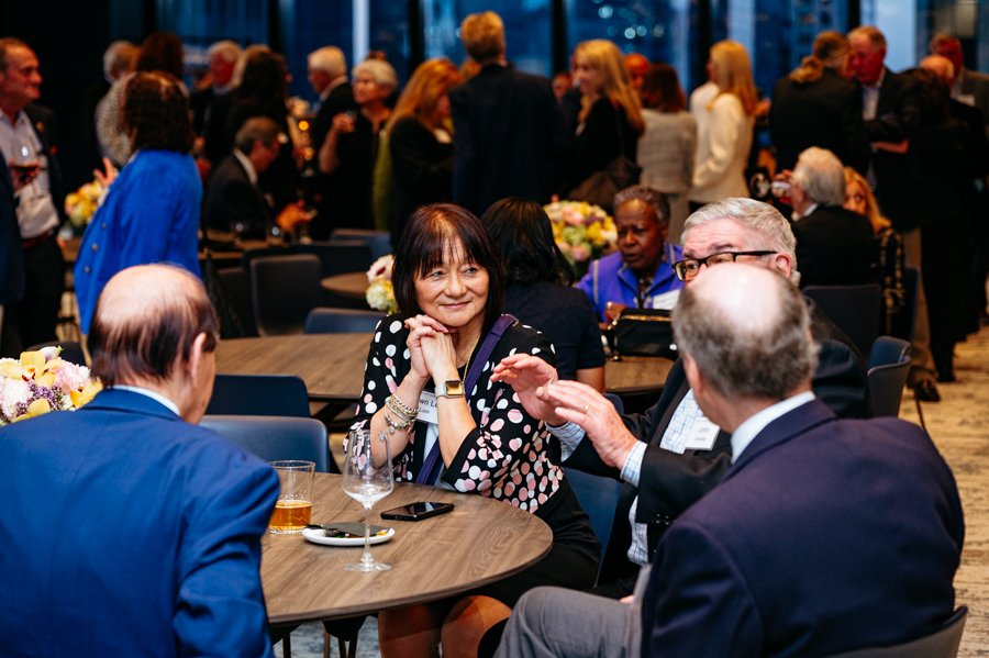 People socializing at an indoor event, some sitting at tables with flowers, others standing and mingling in background.