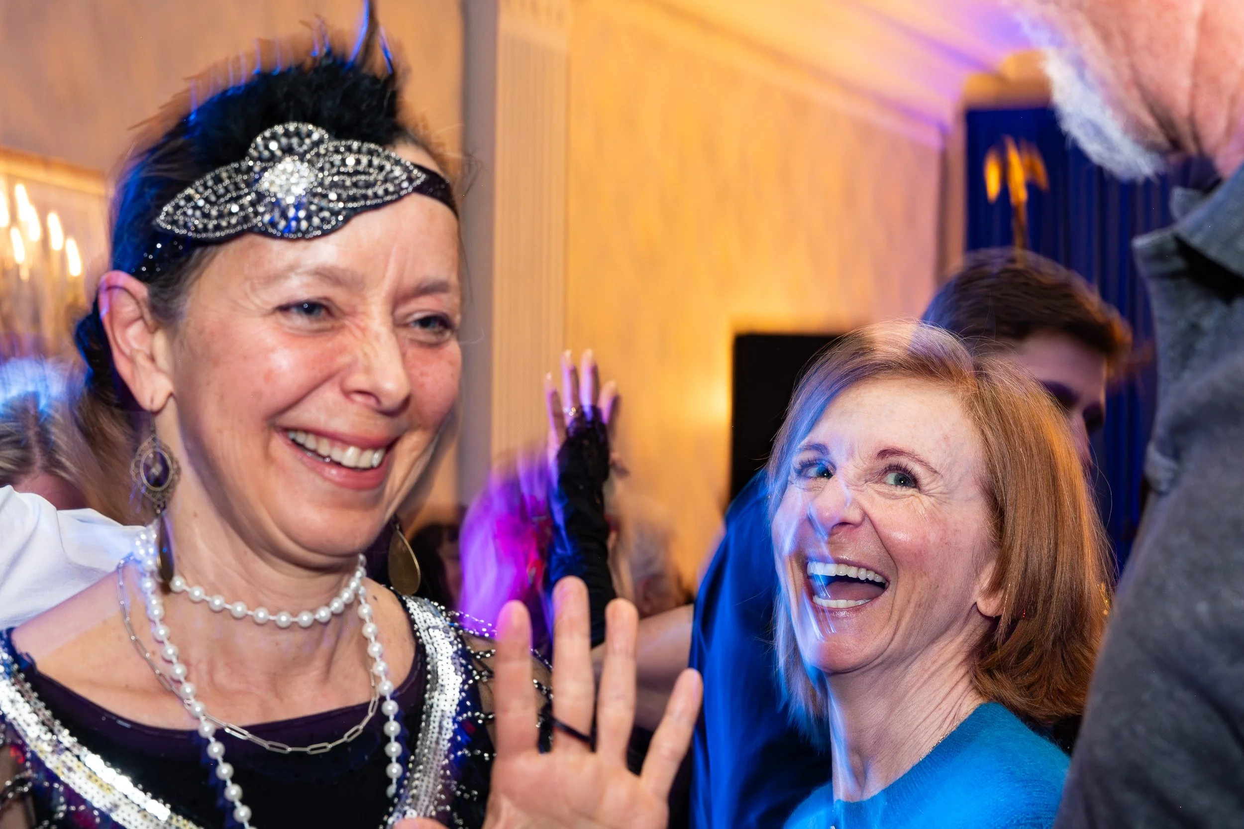Two women smiling and waving at a social gathering, with one wearing a decorative headband and jewelry, and the other wearing a bright blue top.