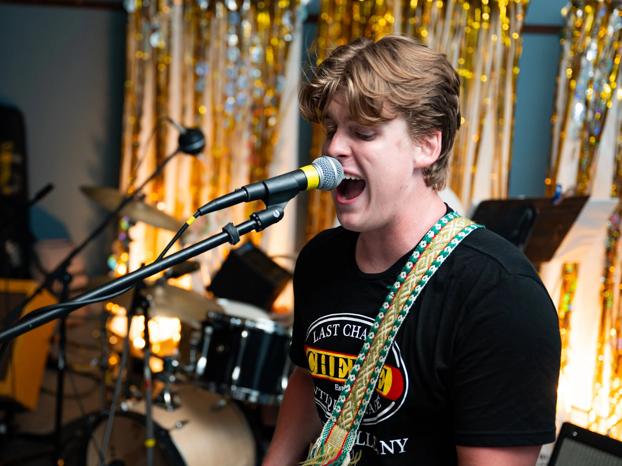 A young man with wavy, light brown hair singing into a microphone during a performance. He is wearing a black t-shirt and a colorful, patterned strap over his shoulder. The background features a curtain decorated with gold tinsel.