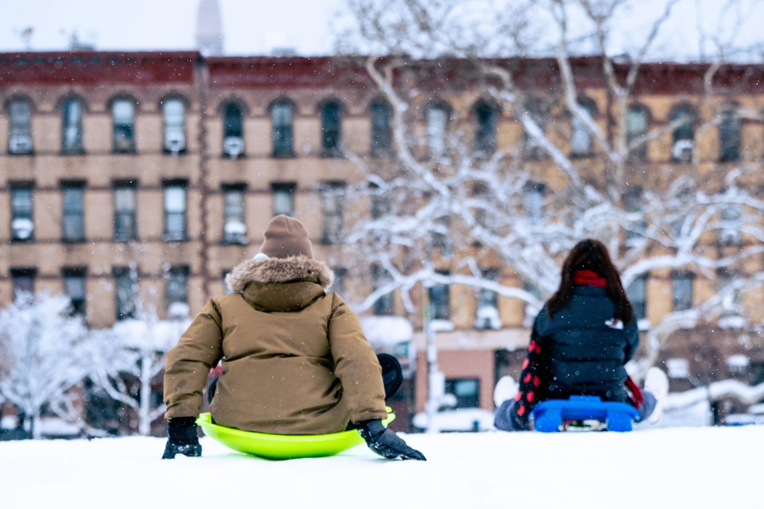 Two children sitting on sleds in snow, facing a building with bare trees in winter.