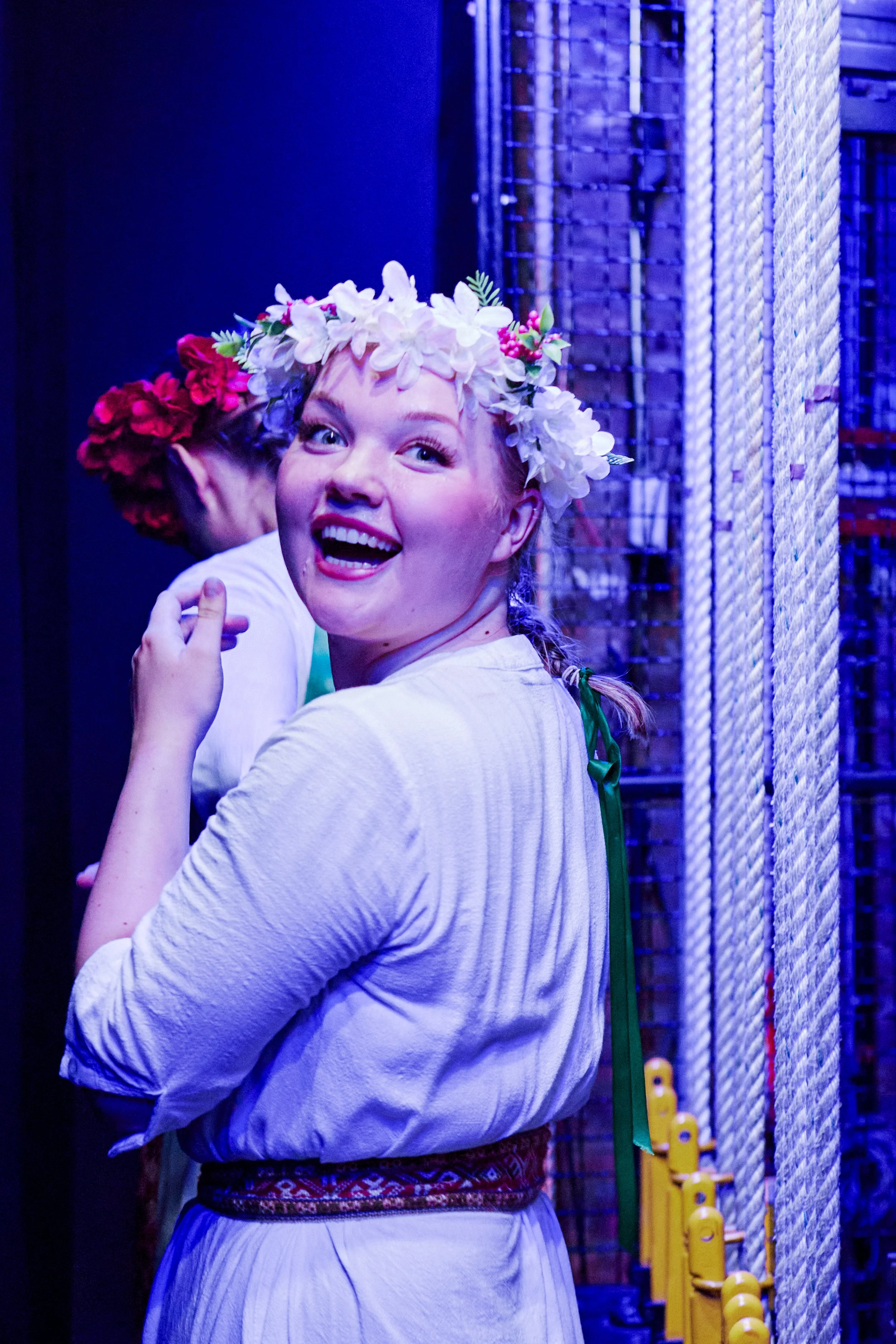 Young woman with a flower crown smiling and turning back, in a colorful, possibly theatrical setting.