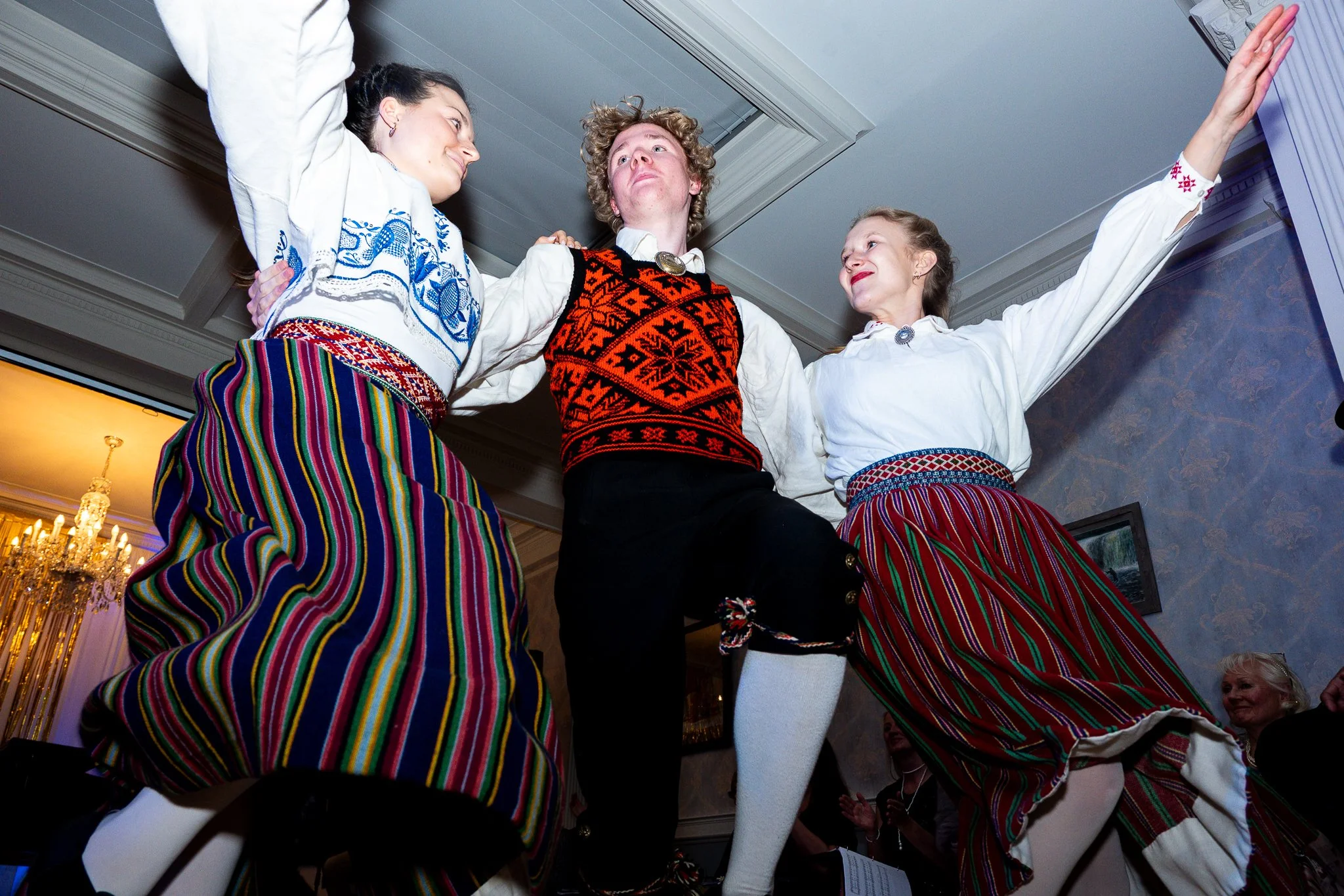 Three people are dancing in traditional clothing indoors. The women are wearing colorful striped skirts and white tops, while the man is dressed in a patterned vest and pants. They are smiling and look joyful.