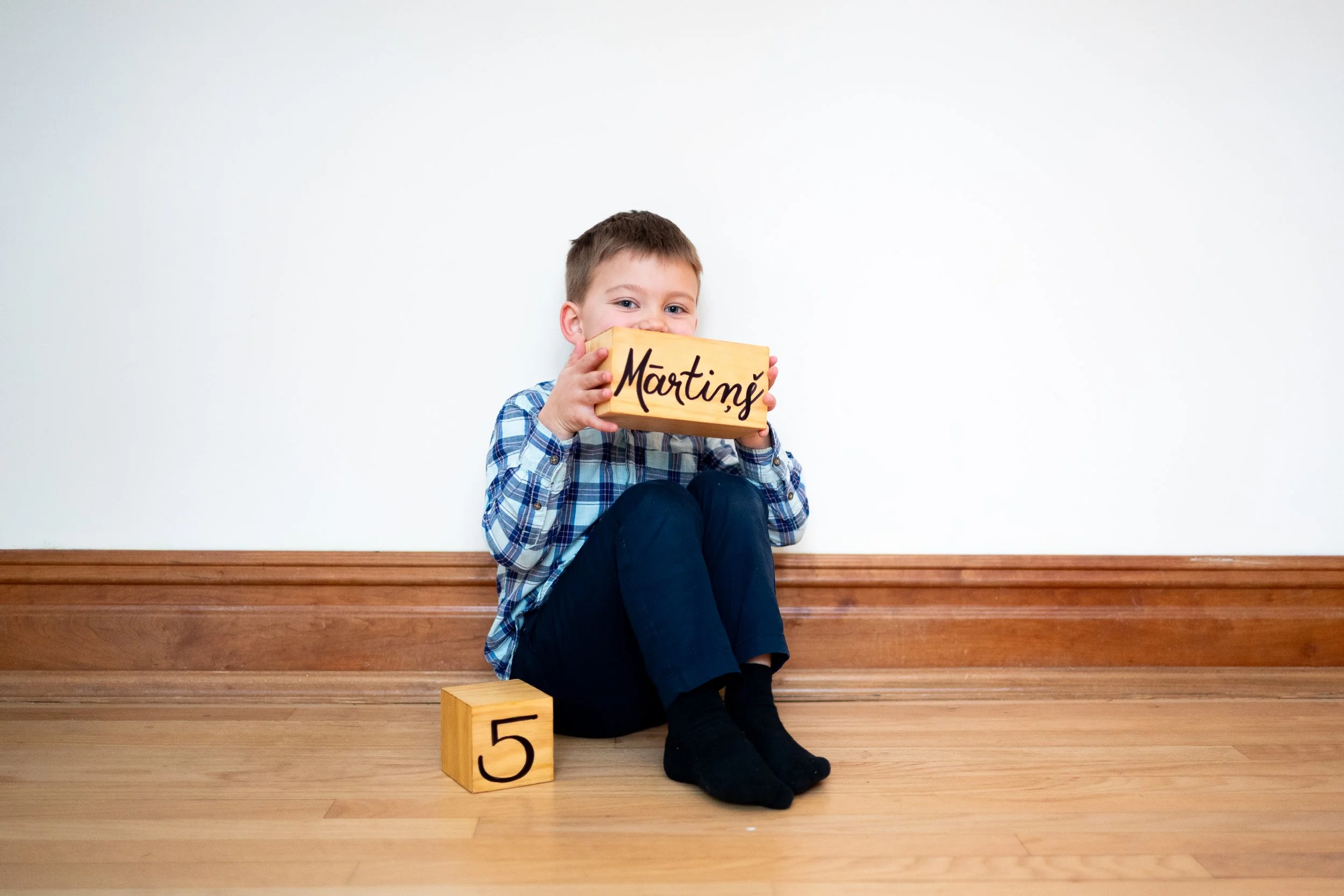 A young boy with short hair and blue eyes sitting on wooden stairs, holding a wooden block with the number 5 and a rectangular wooden sign with the name 'Marting' written on it, smiling at the camera.