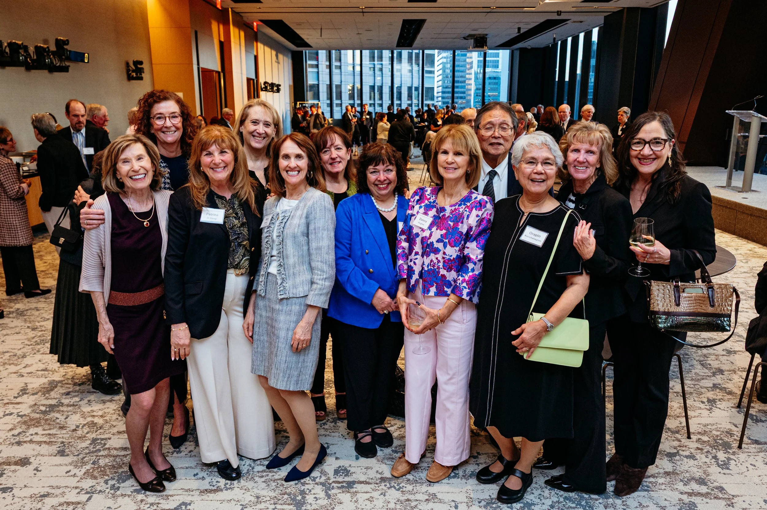 A group of women and one man dressed in business attire, smiling and posing together at a professional event in a modern conference room with large windows showing a cityscape in the background.