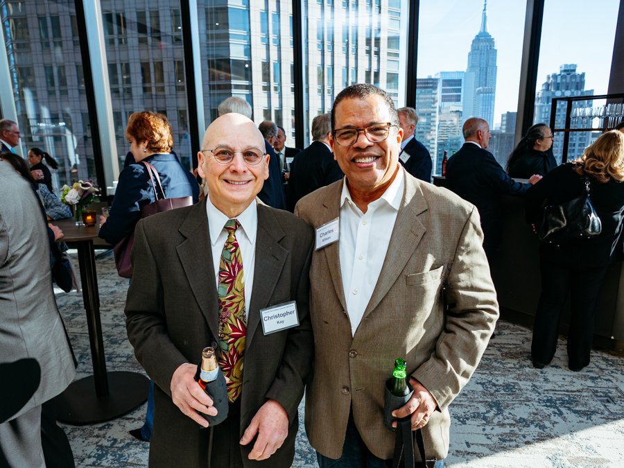 Two men in suits smiling at a social event in a high-rise building with a city skyline in the background, one of the men holding a beer and the other holding a bottle.