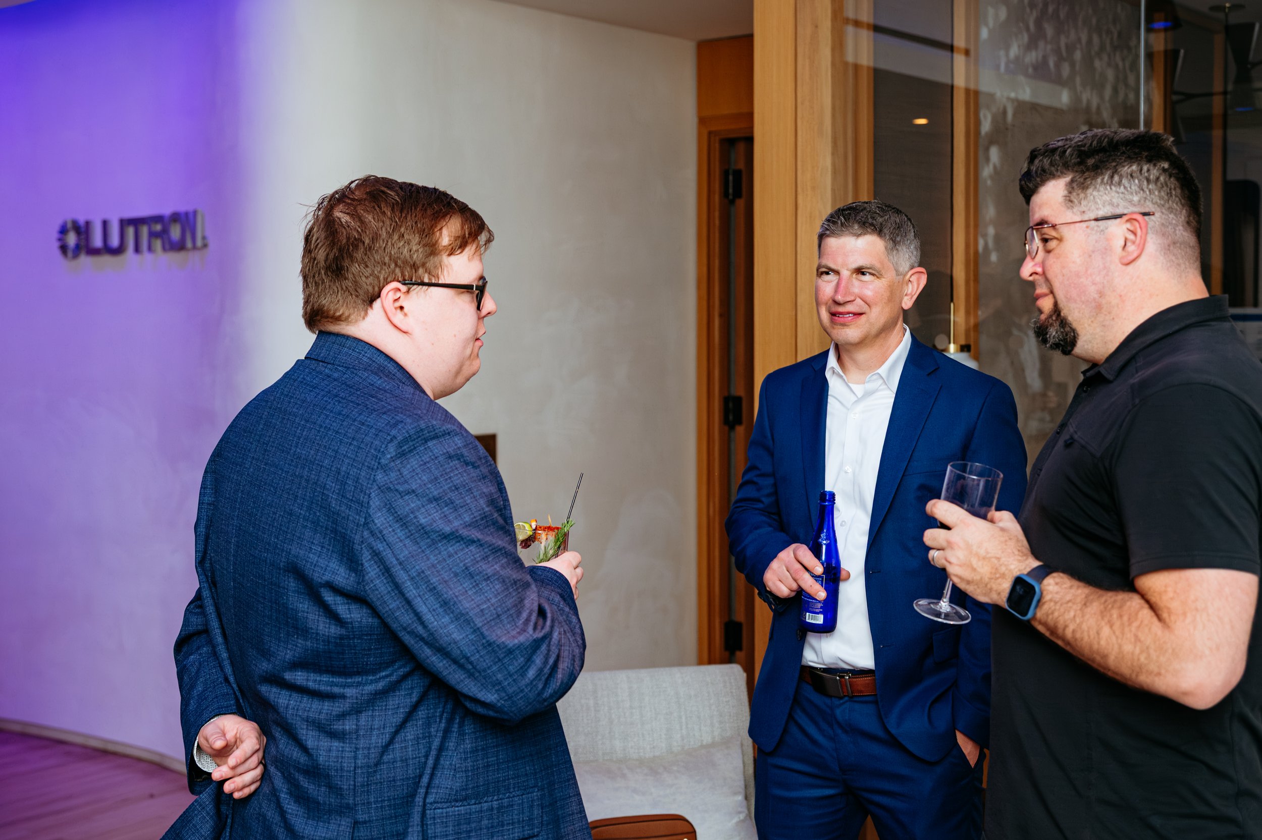 Three men are having a conversation at a social event. They are holding drinks, and one man is wearing a blue suit, another is in a black shirt, and the third is in a blue blazer. There is a purple light and the logo 'LUTRON' on the wall behind them.