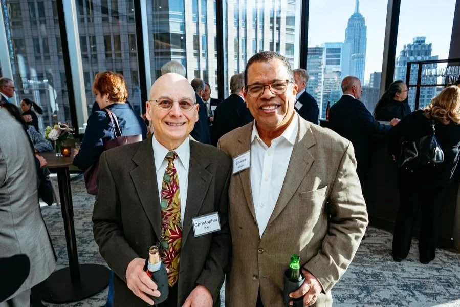 Two smiling men in suit jackets, one with glasses and a floral tie, holding drinks, standing in a high-rise office with large windows and city skyline in the background, at a professional networking event.